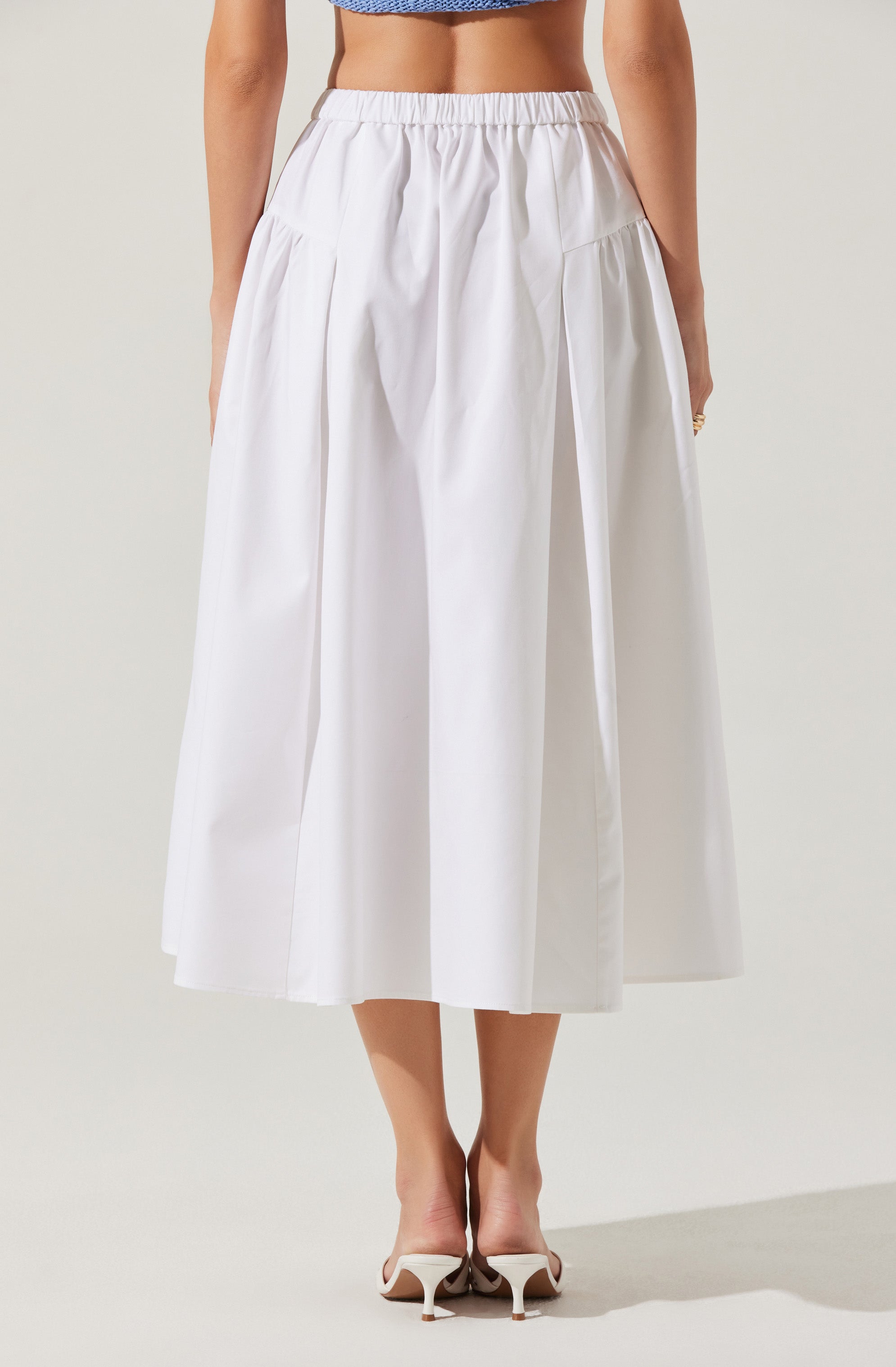 A woman is wearing a high-waisted, white, ankle-length skirt with pleats and a gathered elastic waistband. She is also wearing a blue top that is cropped, revealing her midriff. The skirt has a smooth, tailored appearance, and she is wearing white high-heeled mules.