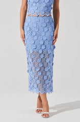 A woman is wearing a matching two-piece outfit made of light blue lace fabric with floral and geometric patterns. The top is sleeveless with a cropped length, and the skirt is high-waisted and reaches mid-calf, featuring scalloped edges at the hem. The lace fabric is semi-sheer, revealing a hint of skin underneath.