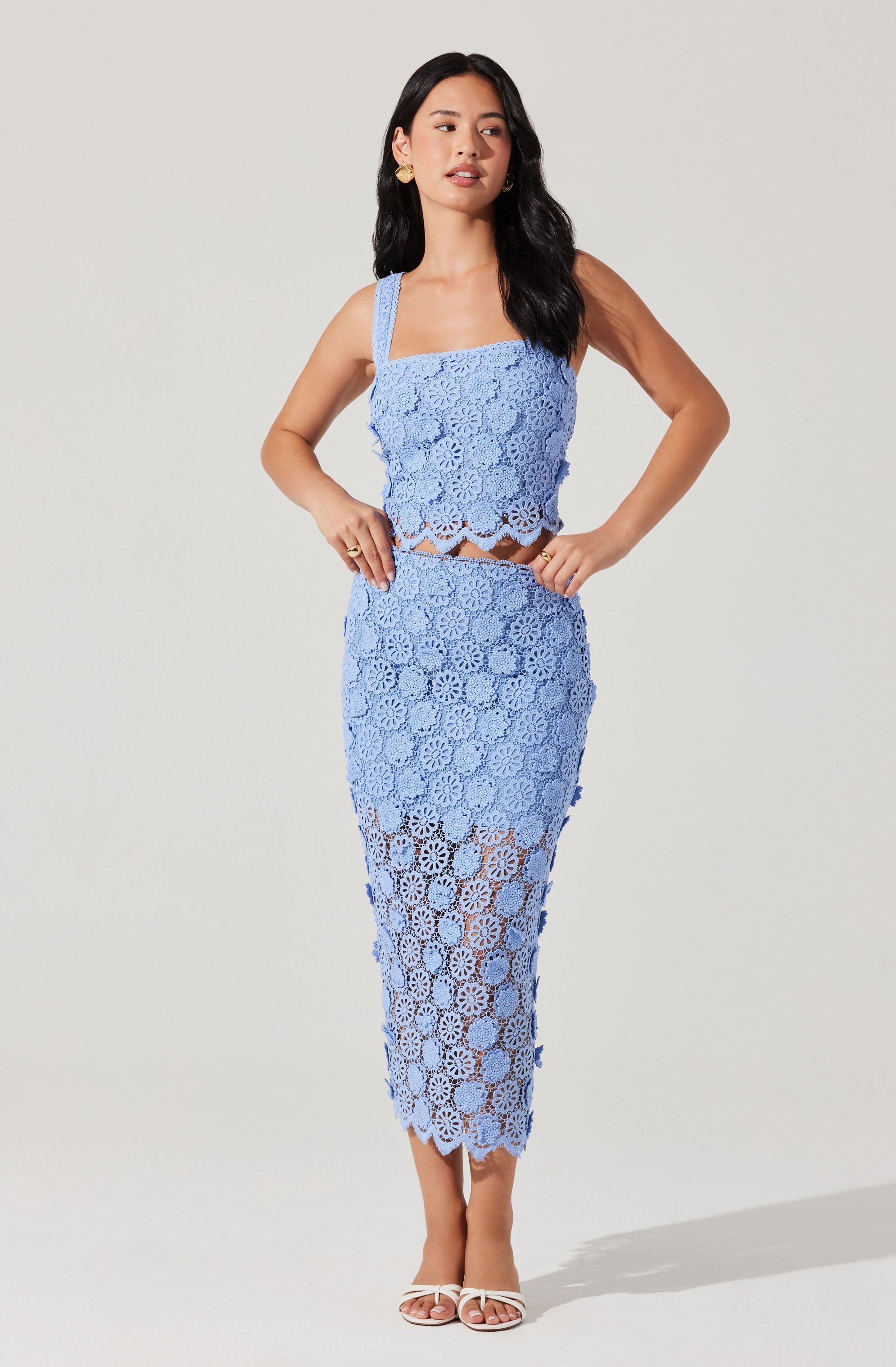 A woman is wearing a matching light blue crochet two-piece outfit consisting of a sleeveless crop top with thick straps and a high-waisted, ankle-length skirt. The crochet pattern features floral motifs throughout the outfit. She is also wearing white high-heeled sandals and gold earrings.