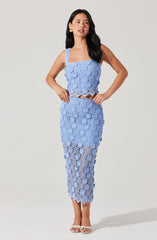 A woman is wearing a matching light blue crochet two-piece outfit consisting of a sleeveless crop top with thick straps and a high-waisted, ankle-length skirt. The crochet pattern features floral motifs throughout the outfit. She is also wearing white high-heeled sandals and gold earrings.