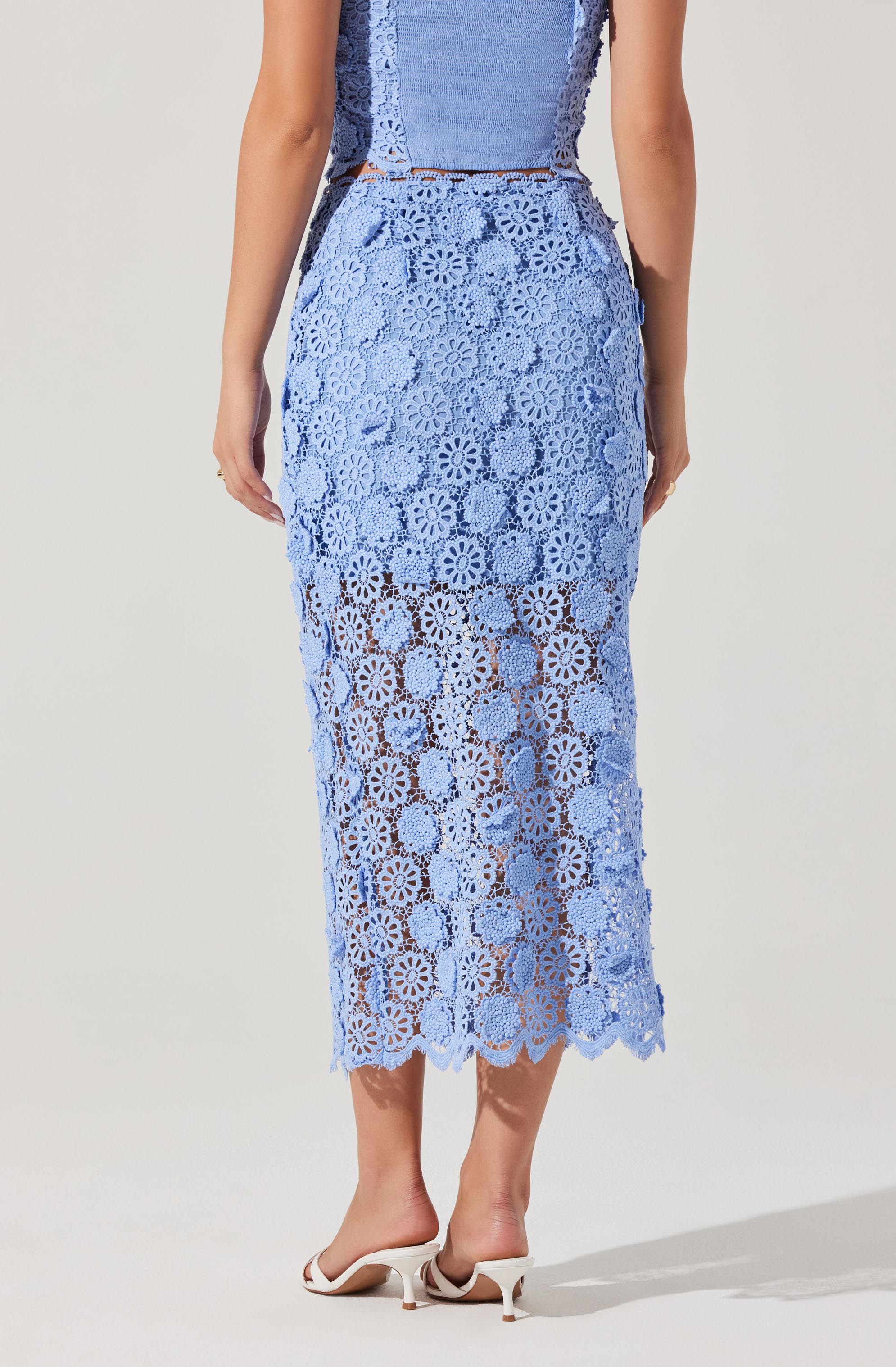 A woman is wearing a light blue lace dress with a floral pattern. The dress has intricate floral lace detailing, and the hemline falls below the knees. The dress is semi-sheer with a lining underneath in the upper part and transparent lace pattern towards the bottom. She is also wearing white heeled sandals.