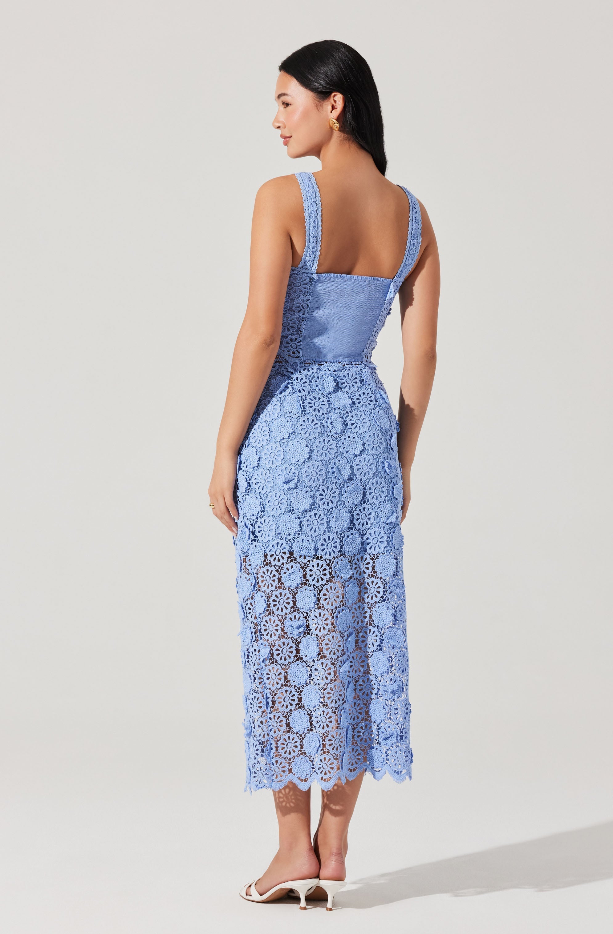 A woman is wearing a sleeveless, light blue crochet dress with a fitted bodice and wide shoulder straps. The dress features intricate crochet patterns throughout, with the skirt portion being semi-sheer and extending to mid-calf length. She is also wearing white heeled sandals.