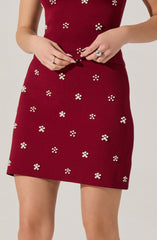 A woman is wearing a red dress decorated with small, white, floral-shaped embellishments. The dress is fitted at the waist and has a straight skirt that falls above the knees. She is adjusting the hem of the dress with her hands.