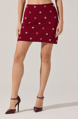 A woman is wearing a high-waisted, burgundy mini skirt decorated with small, white flower patterns scattered throughout. She is also wearing burgundy high-heeled shoes with ankle straps.