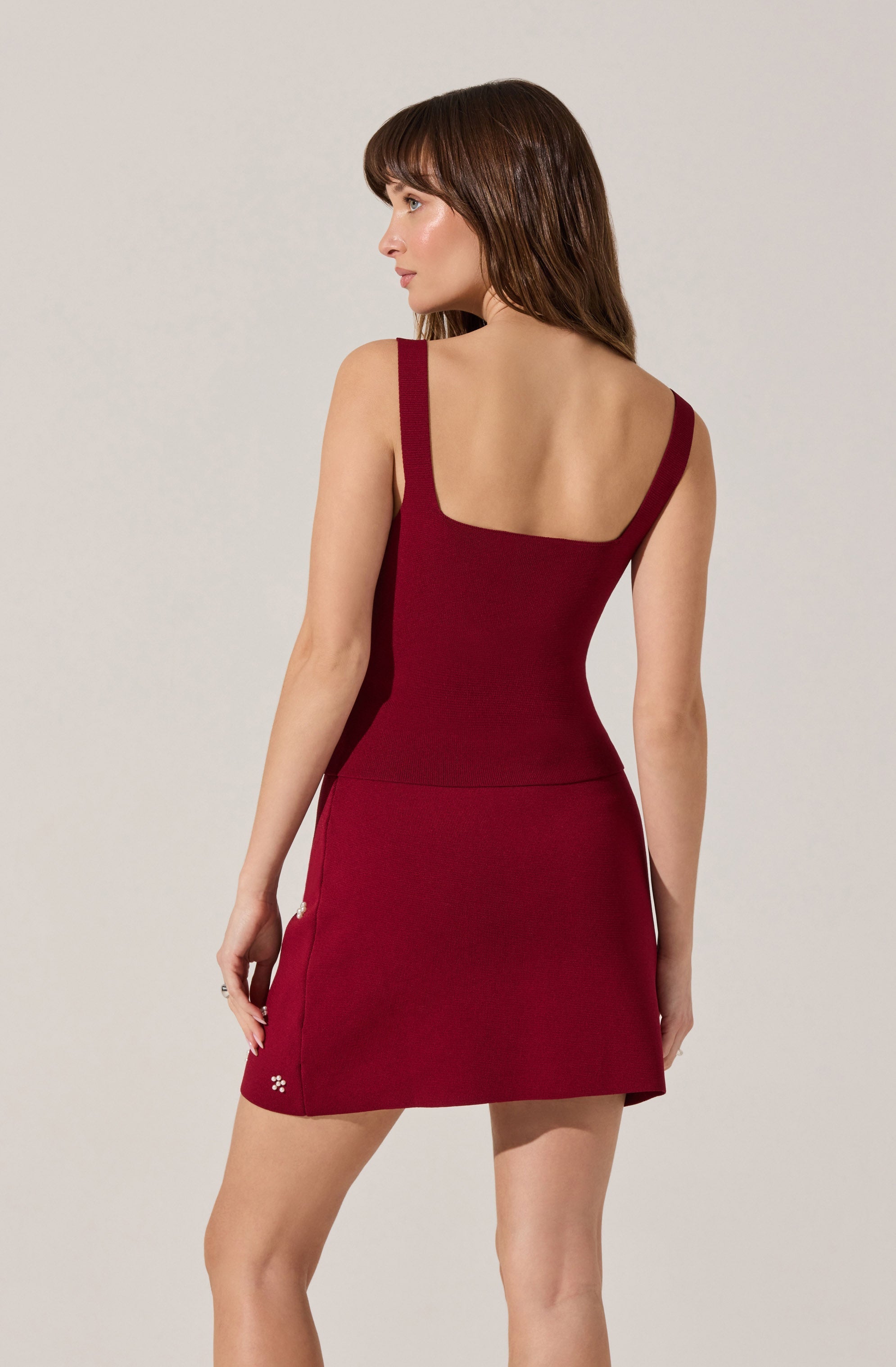 A woman is wearing a sleeveless, fitted dress with wide shoulder straps and a scoop neckline. The dress is deep red in color and has a smooth, streamlined design.