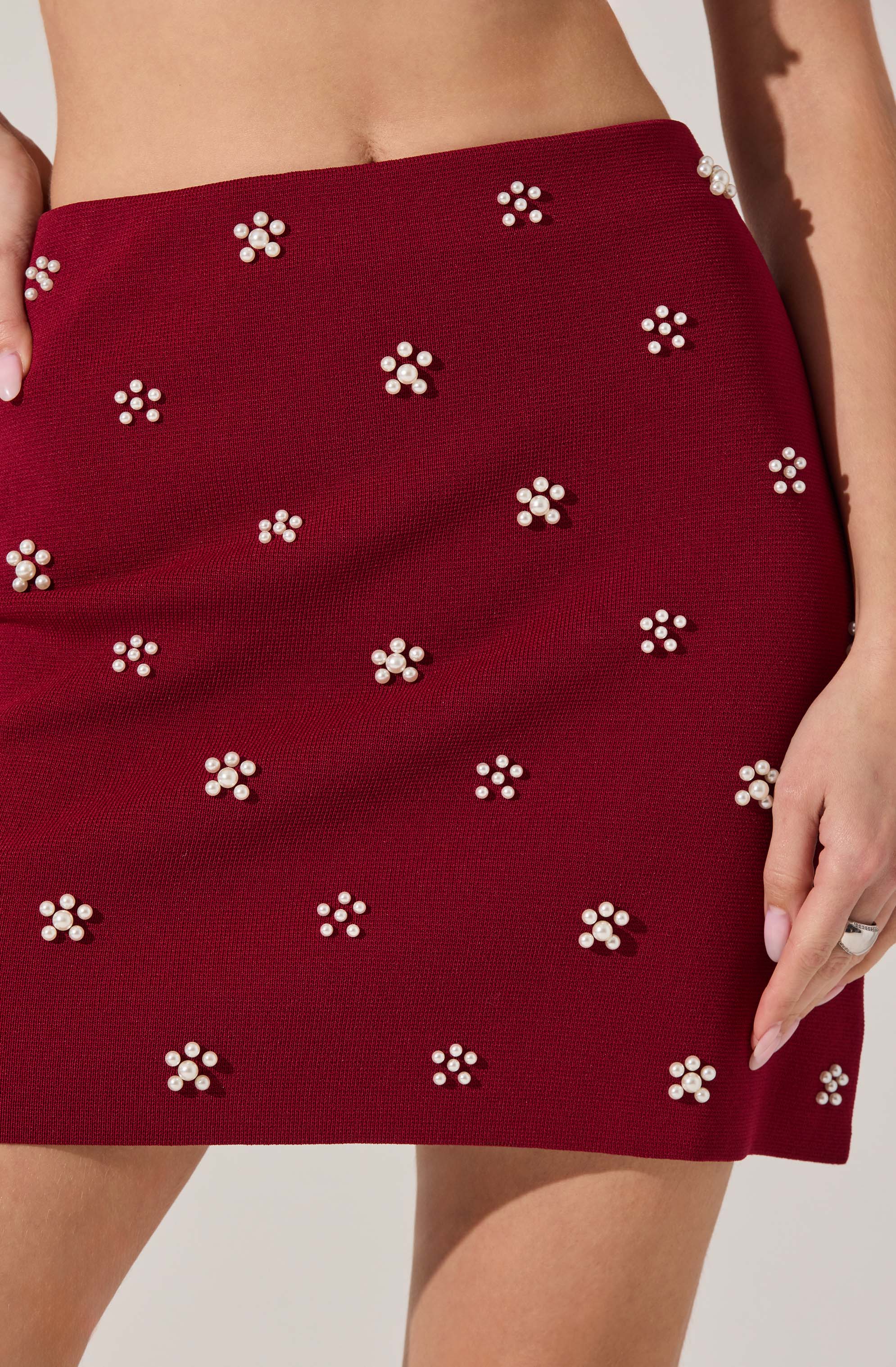 A close-up of a woman's red skirt decorated with small clusters of white pearls arranged in flower shapes. The skirt has a textured fabric and sits high on the waist. The woman's left hand rests on her thigh, with one finger slightly touching the fabric.
