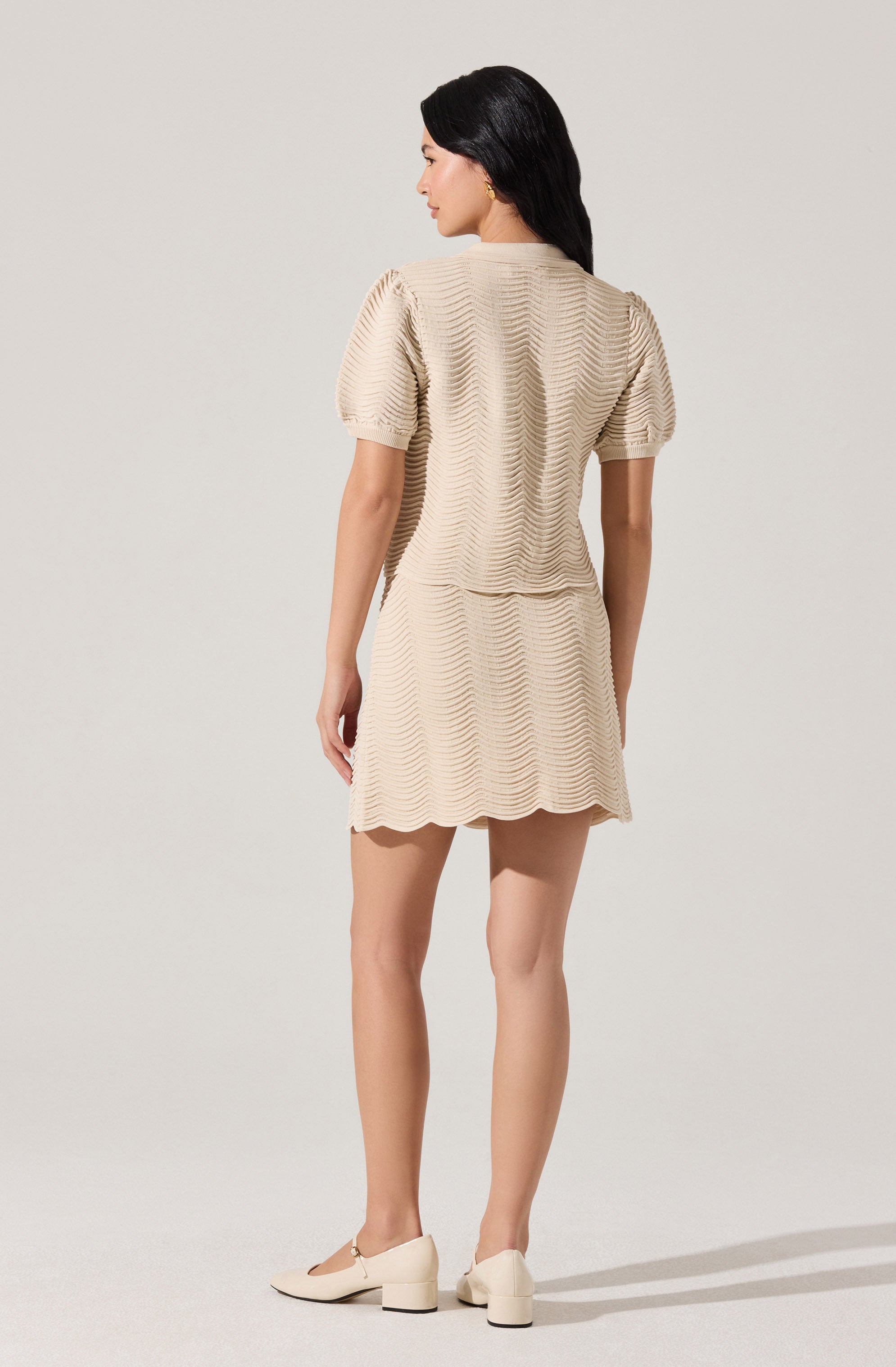 A woman is wearing a beige, textured dress with a subtle wave pattern. The dress has puffed short sleeves and a slightly flared skirt that falls above the knees. She is also wearing cream-colored shoes with a small heel.