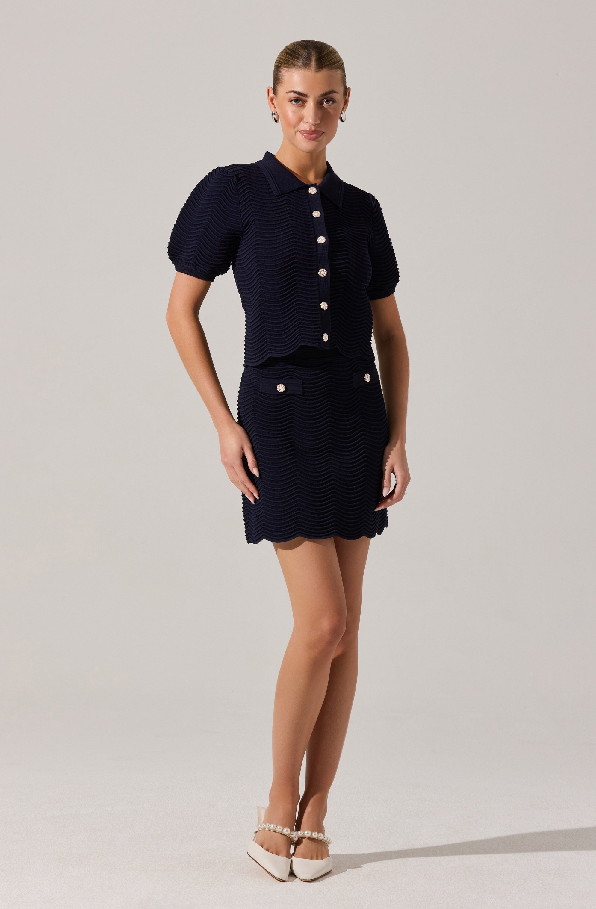 A woman is wearing a matching navy blue textured short-sleeve button-up top with a collar and a matching textured skirt with white buttons and pockets. She pairs the outfit with beige heels decorated with pearl embellishments.