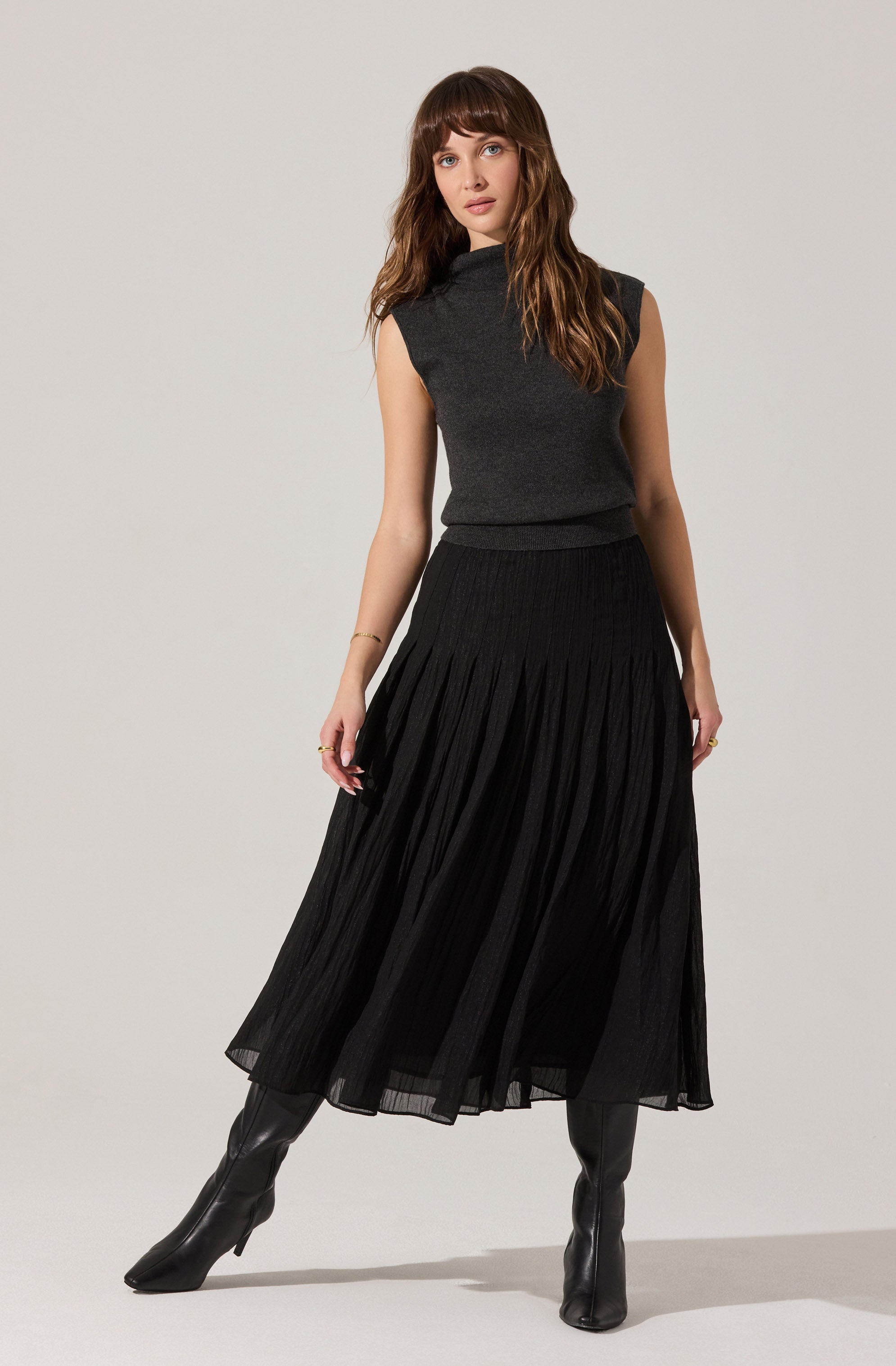 A woman is wearing a sleeveless, dark gray turtleneck top with a slightly loose fit. She pairs it with a long, black pleated skirt that reaches her ankles. She completes her outfit with knee-high black heeled boots.