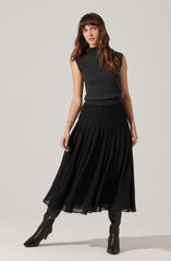 A woman is wearing a sleeveless, dark gray turtleneck top with a slightly loose fit. She pairs it with a long, black pleated skirt that reaches her ankles. She completes her outfit with knee-high black heeled boots.