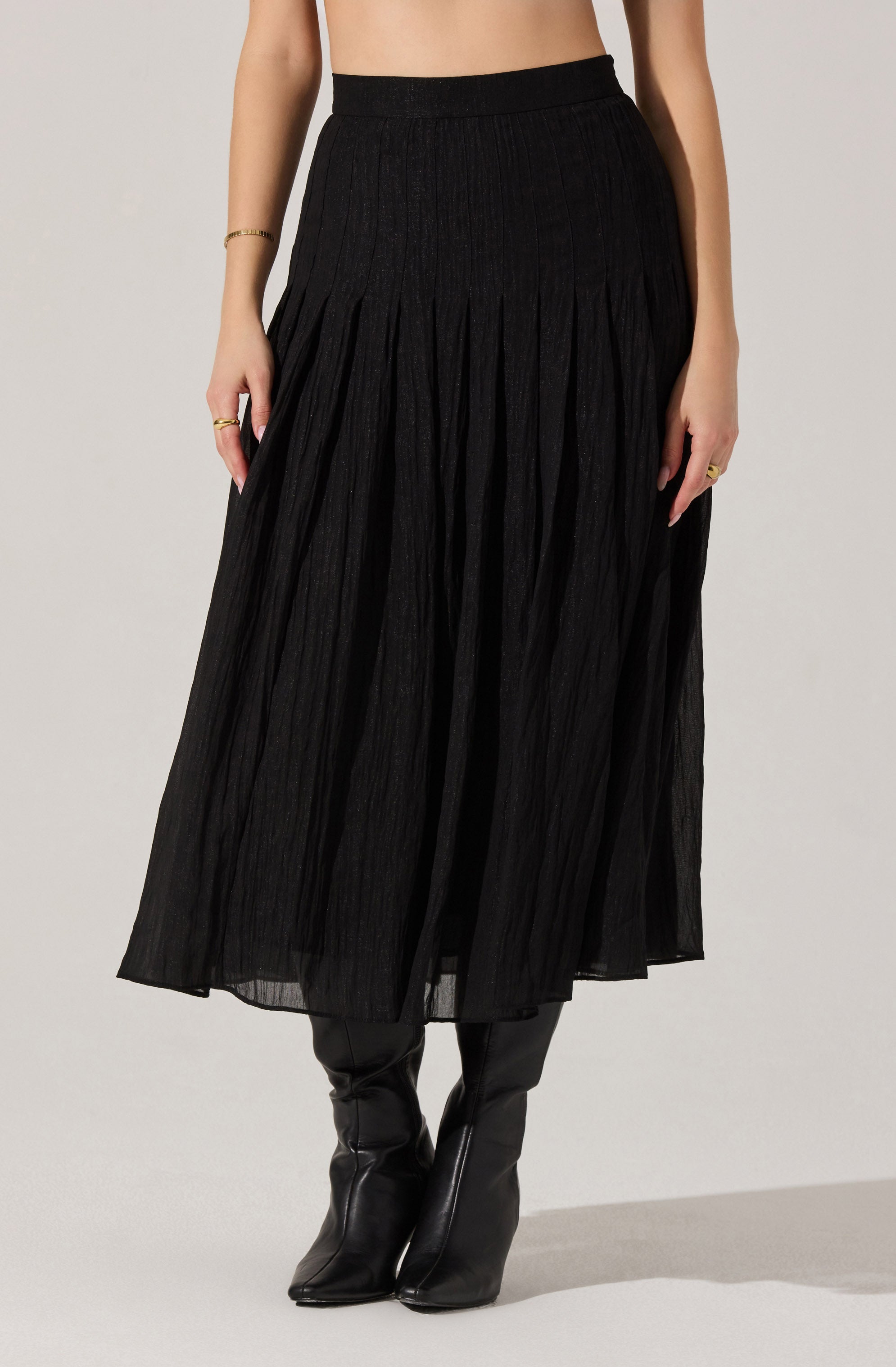 A woman is wearing a high-waisted, black, pleated midi skirt made of lightweight, semi-sheer fabric. She pairs the skirt with black pointed-toe boots. She is also wearing a delicate bracelet and rings on her hands.