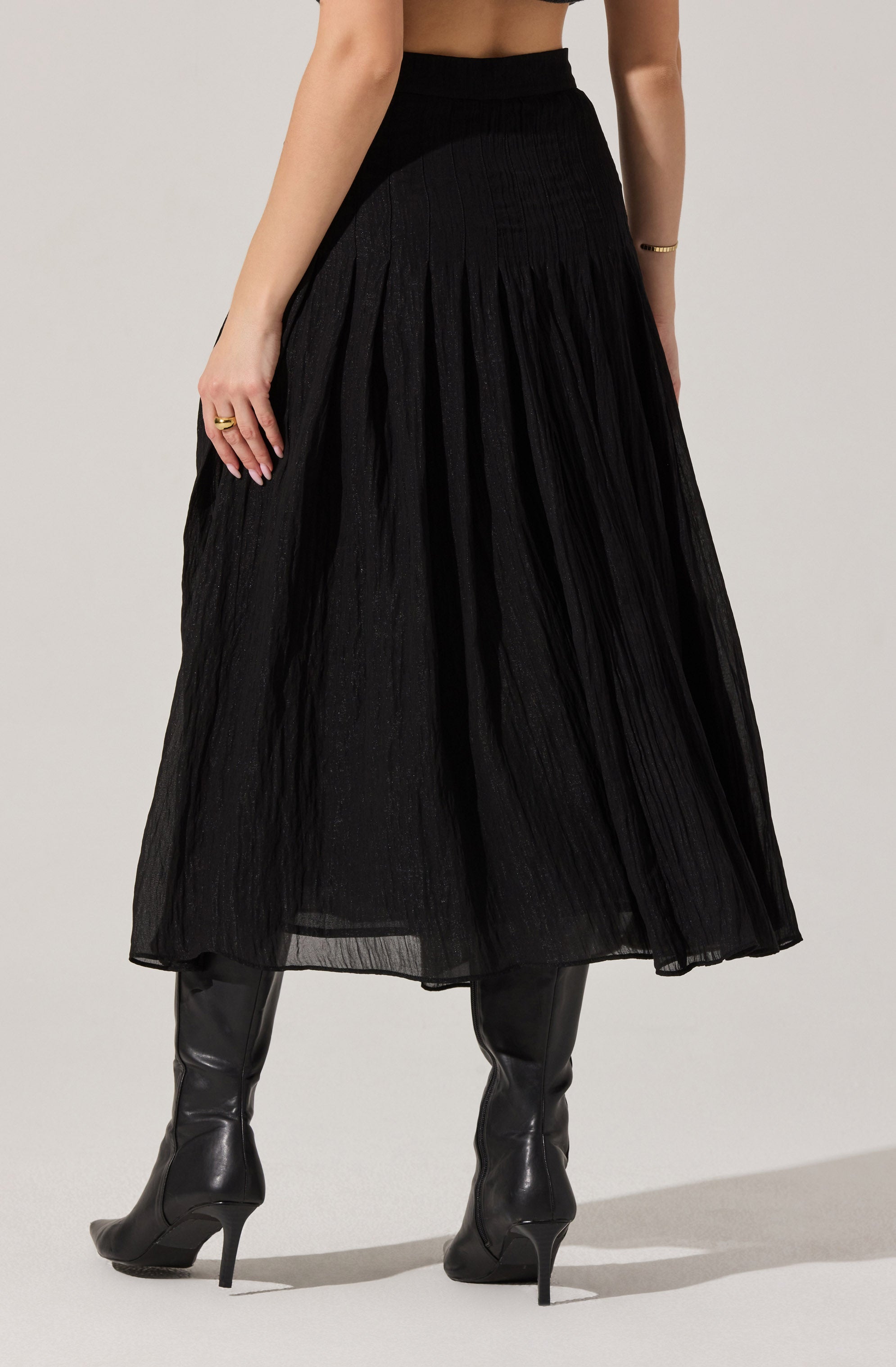 The outfit features a high-waisted, ankle-length black skirt with a textured, pleated design. The model is also wearing black knee-high heeled boots made of shiny leather material.