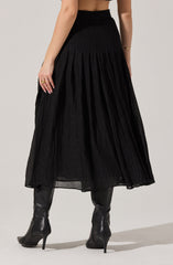 The outfit features a high-waisted, ankle-length black skirt with a textured, pleated design. The model is also wearing black knee-high heeled boots made of shiny leather material.