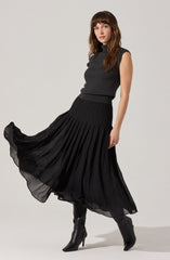 A woman is wearing a sleeveless, high-neck, dark gray top and a long, flowing black pleated skirt. She has on black heeled boots.