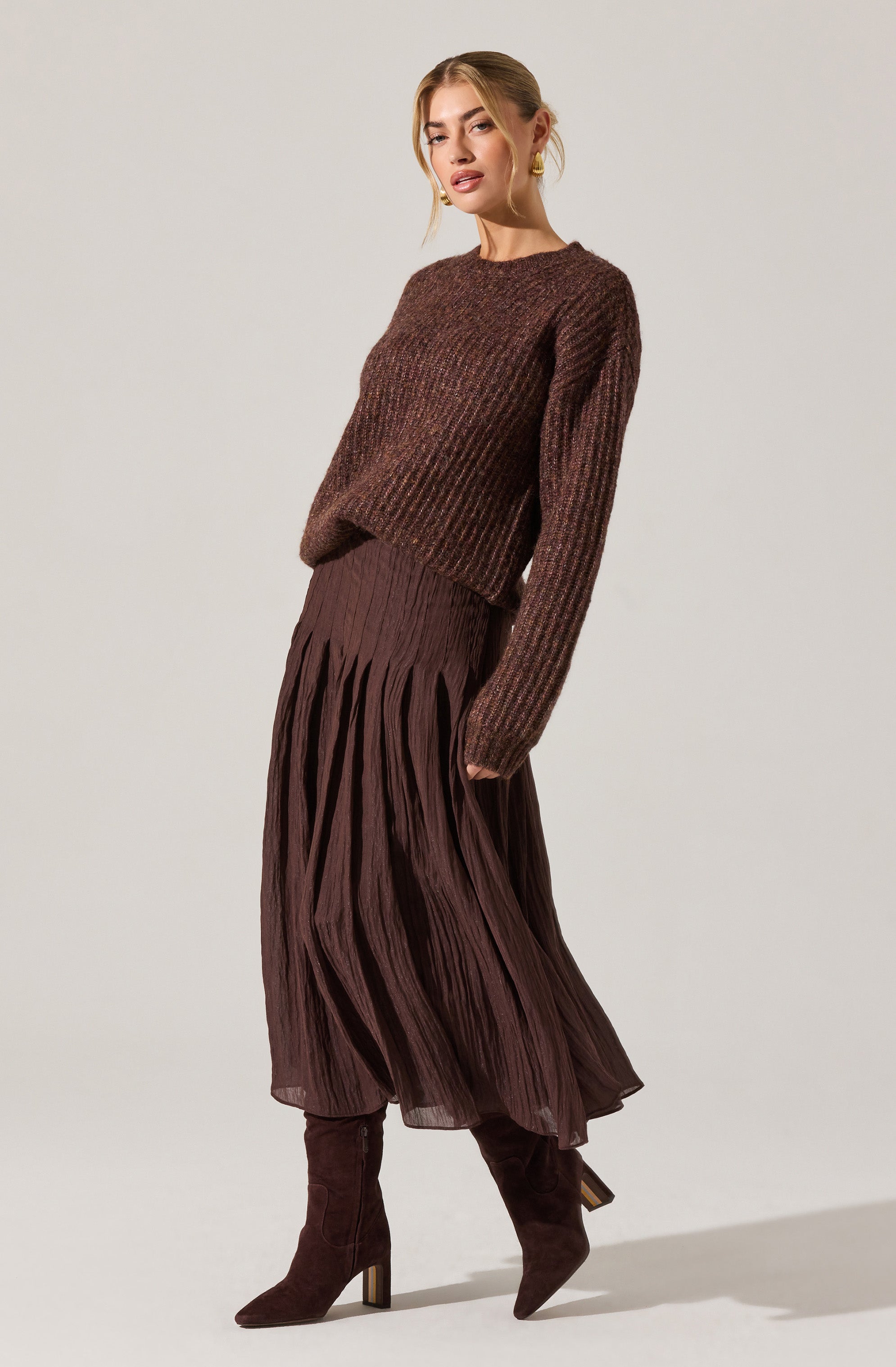 A woman is wearing a brown knit sweater with a textured pattern, long sleeves, and a round neckline. She pairs it with a high-waisted, midi-length skirt made of pleated, lightweight fabric in a matching brown color. She completes the outfit with dark brown suede high-heeled boots that reach just below the knees.