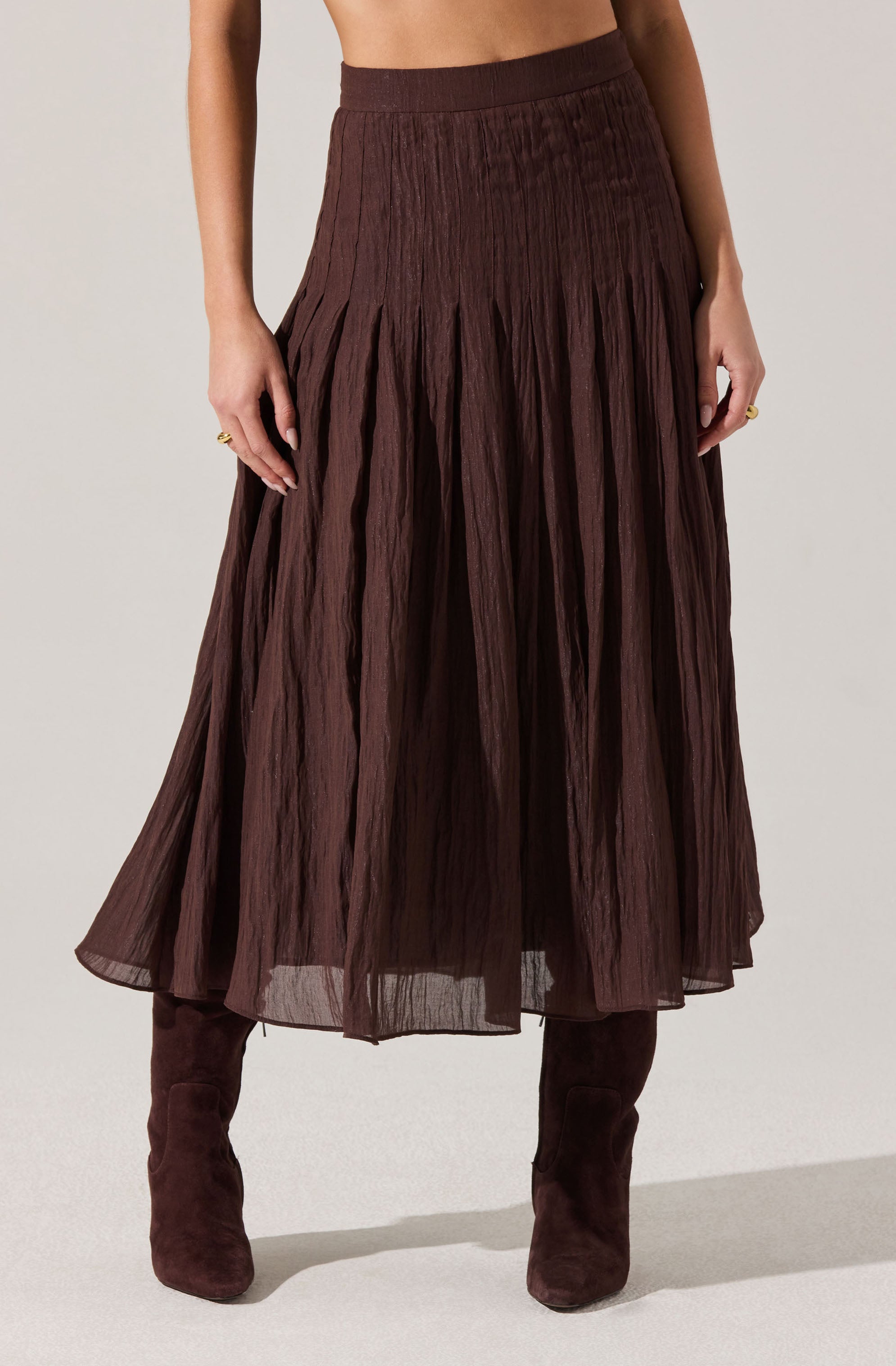 A woman is wearing a high-waisted, pleated, dark brown skirt made from a lightweight, semi-sheer fabric. She has paired it with matching knee-high boots. The skirt has a textured, crinkled appearance with wide pleats that create a flowing silhouette.