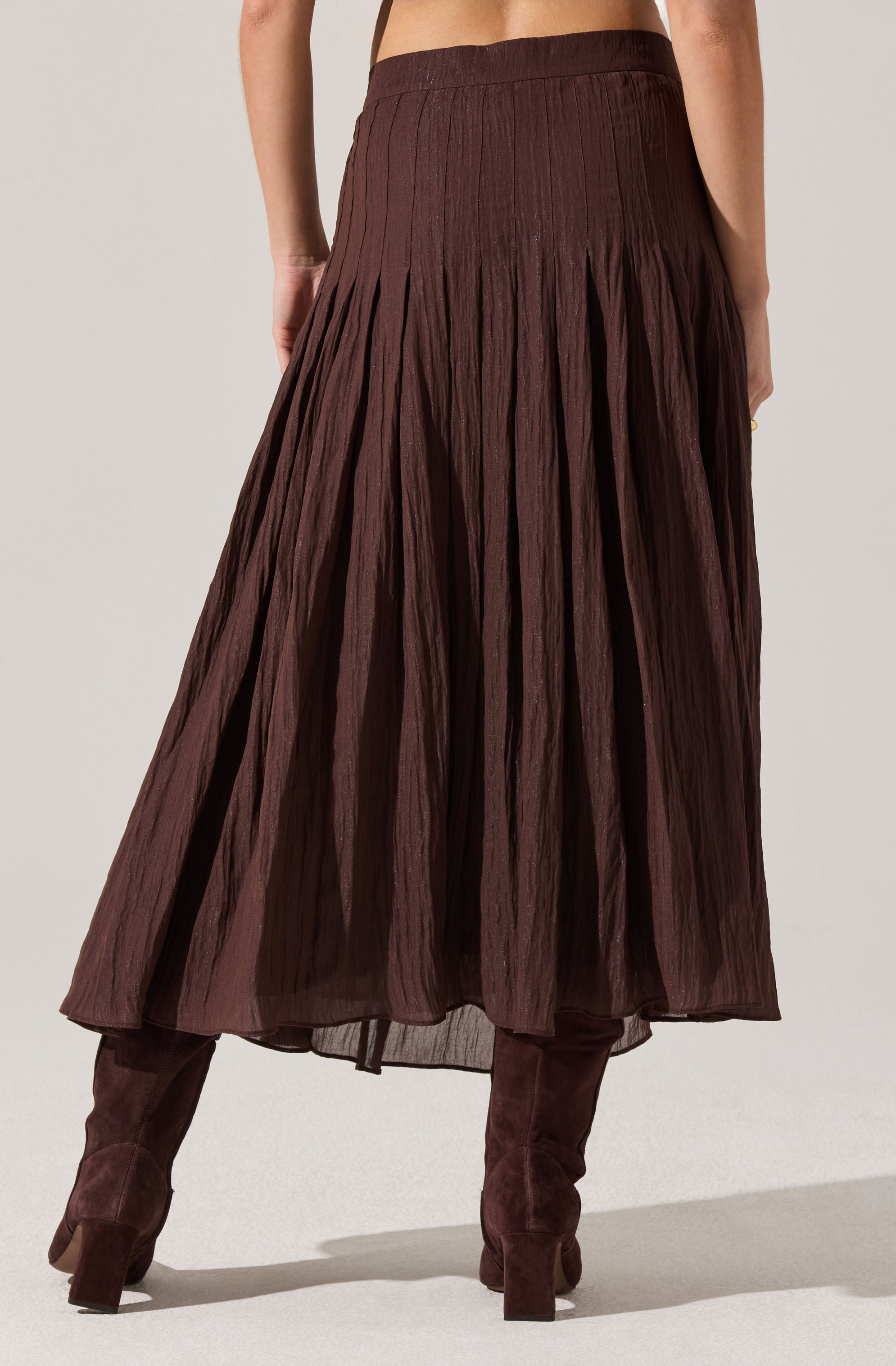 A woman is wearing a long, dark brown pleated skirt paired with matching dark brown knee-high boots. The skirt has a high waistband and a textured, crinkled appearance.