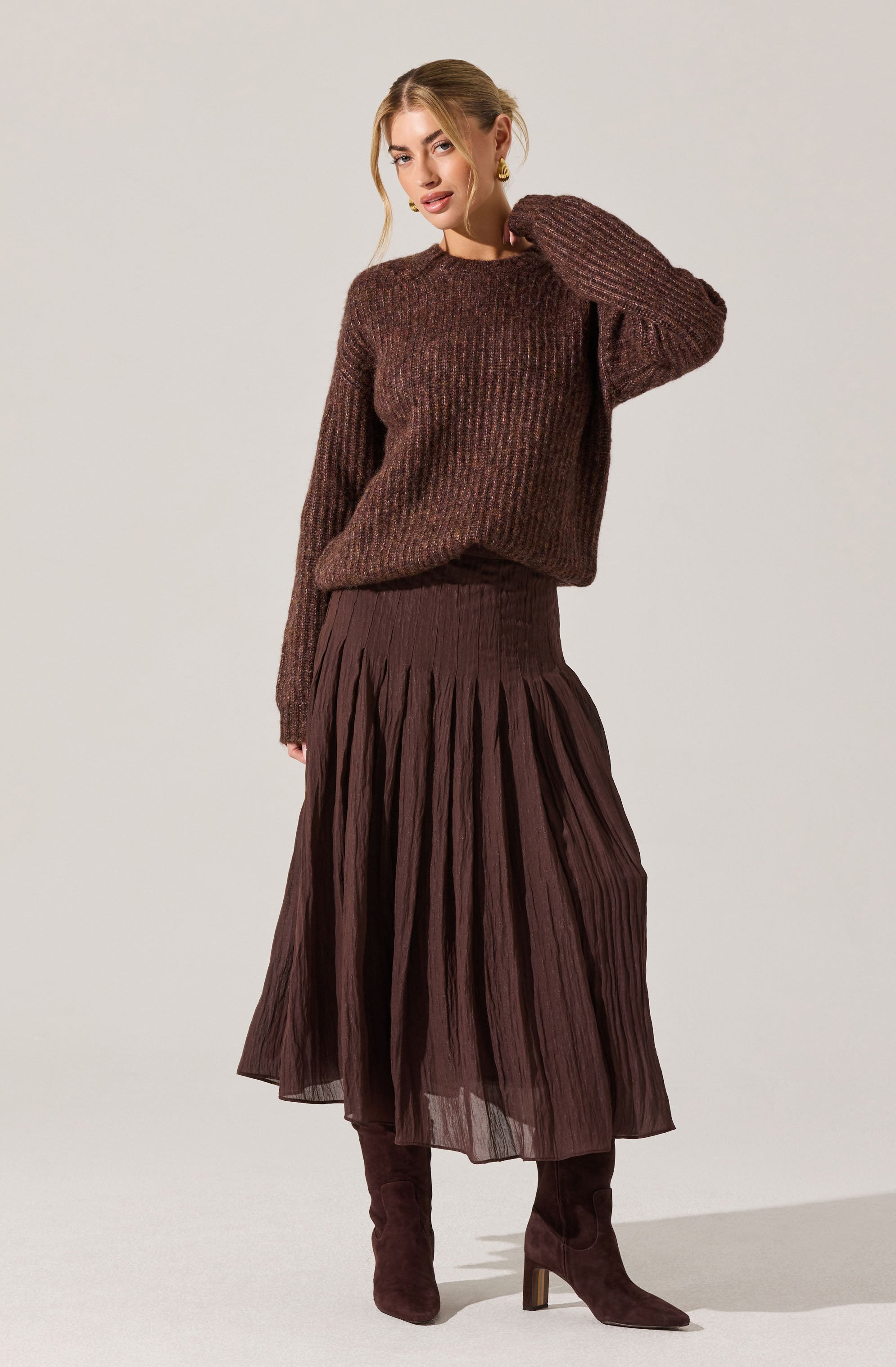 A woman is wearing a chunky, textured, long-sleeved, brown knit sweater with a relaxed fit. She pairs it with a high-waisted, dark brown pleated midi skirt that has a sheer, flowing appearance. She completes her outfit with matching dark brown knee-high boots with a block heel.