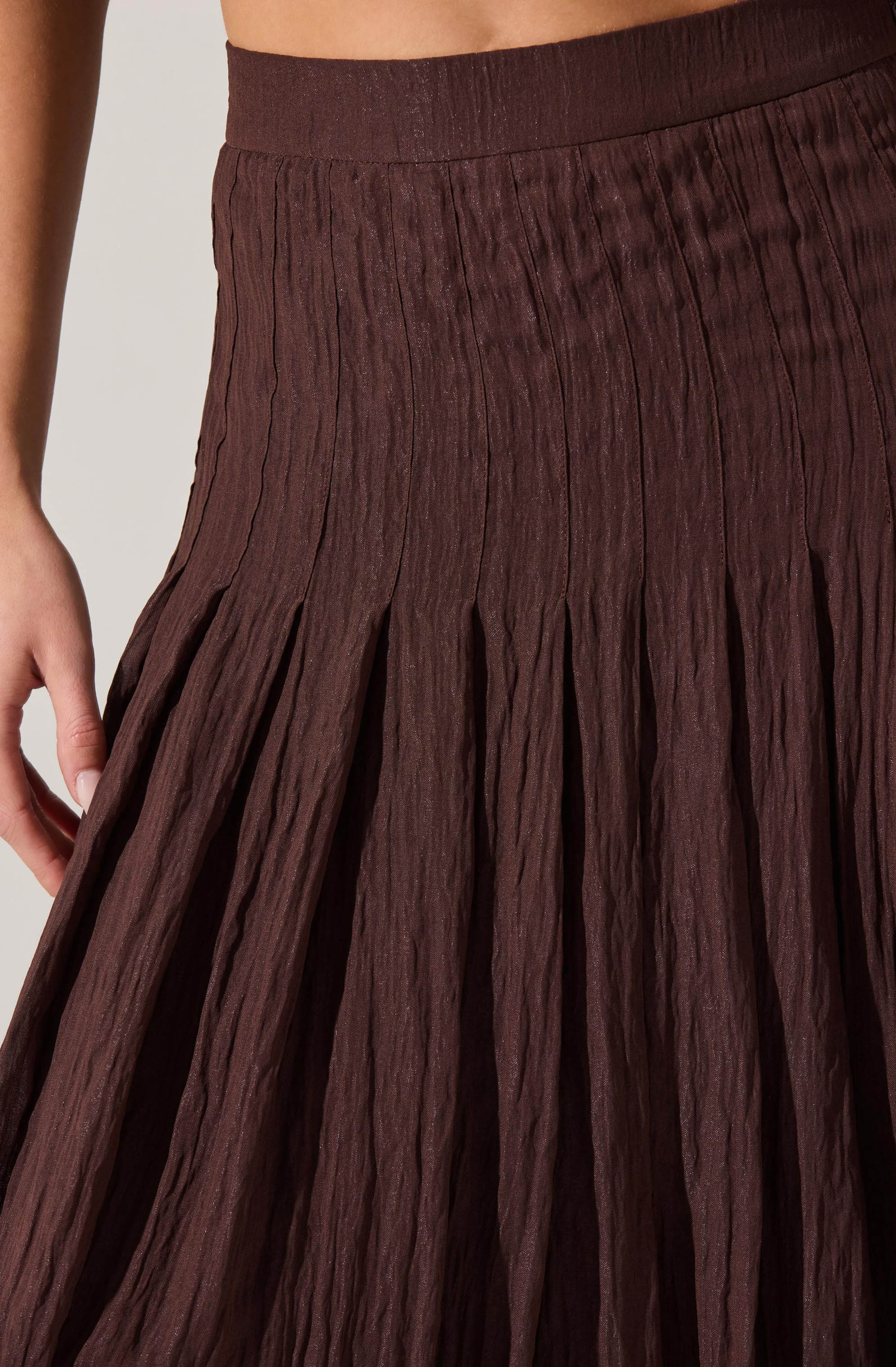 A close-up of a woman's waist and part of her hand, showing a high-waisted skirt made of textured, crinkled fabric in a rich chocolate brown color. The skirt features a wide elastic waistband and pleats that create a flowing, gathered appearance.