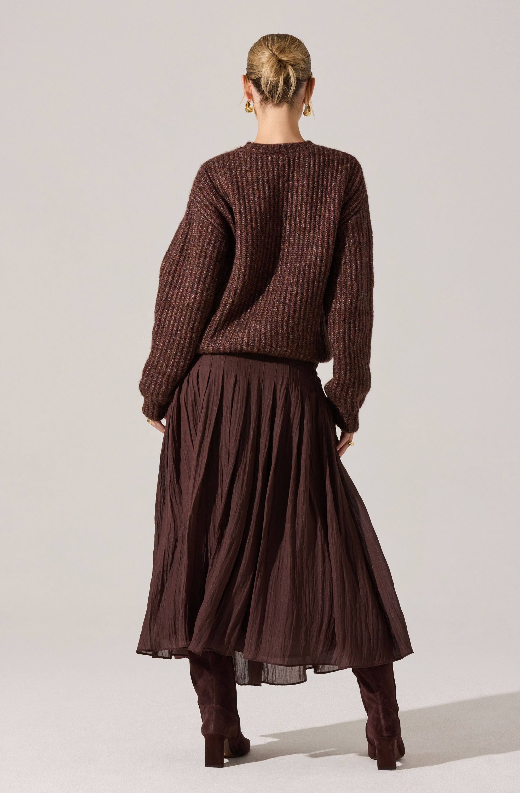 A person is wearing a chunky, ribbed sweater in shades of brown and maroon, paired with a pleated, ankle-length skirt in a deep brown color. They are also wearing matching brown, high-heeled knee-high boots. Their hair is styled in a low bun and they are accessorized with earrings.