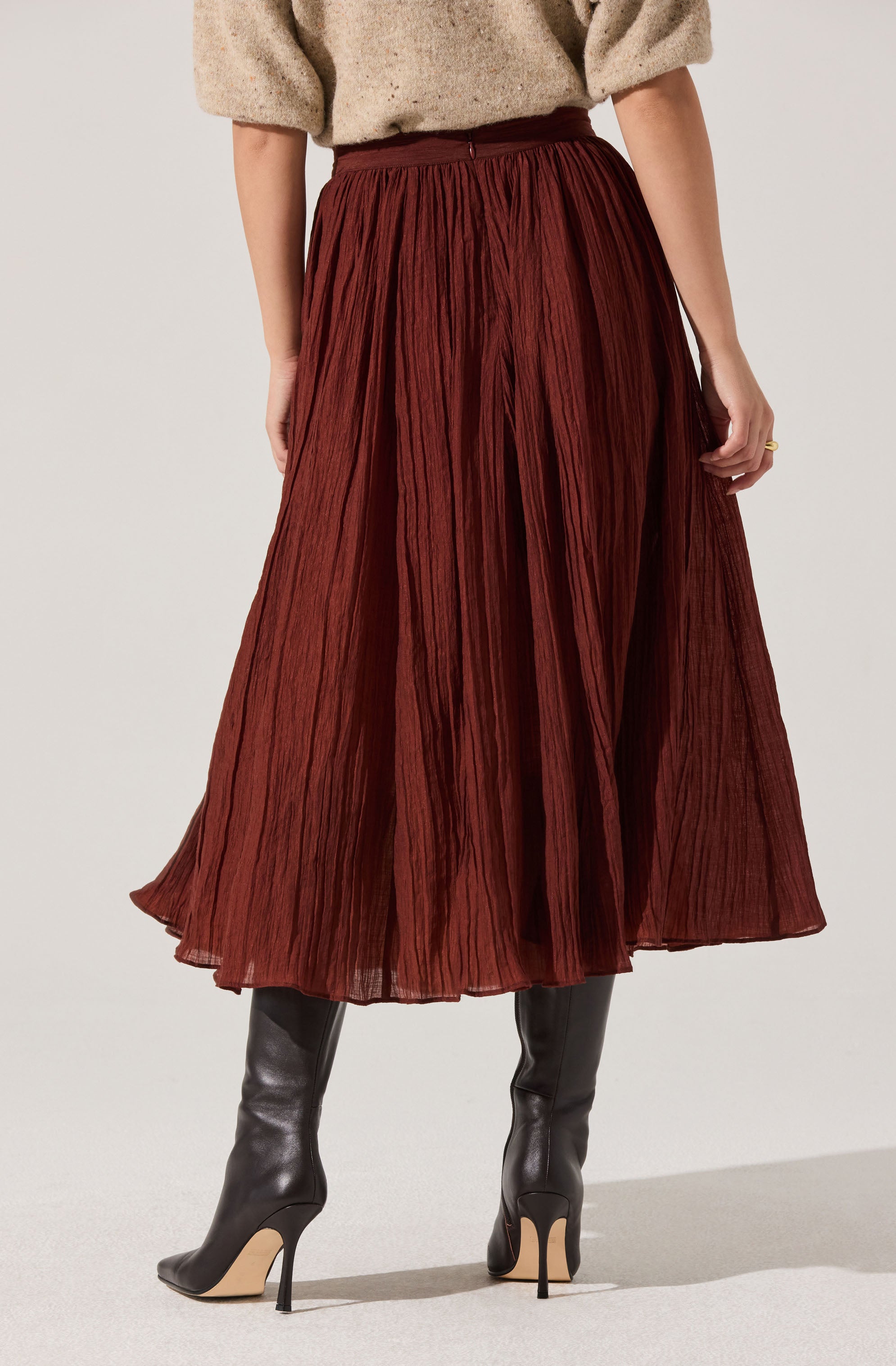 A woman is wearing a high-waisted, ankle-length, pleated skirt made of a textured, rust-colored fabric. She pairs the skirt with a loose, beige sweater and black high-heeled boots.