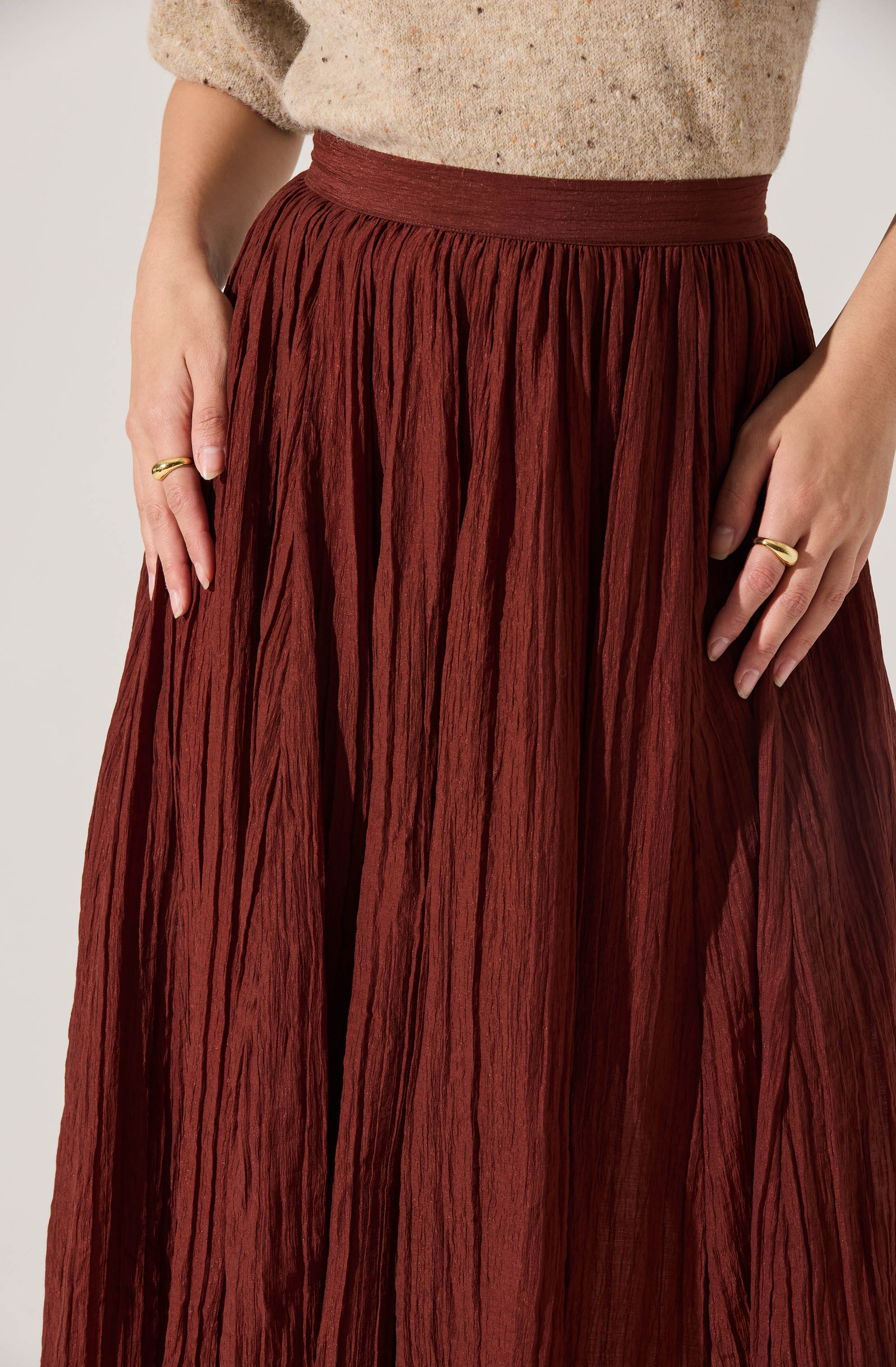 A person is wearing a high-waisted, ankle-length skirt made of crinkled, textured fabric in a rich rust color. The skirt has a wide waistband and vertical pleats that create a flowy silhouette. The person is also wearing a speckled beige top with slightly puffed sleeves. They have both hands resting at their sides, each with a gold ring on their fingers.