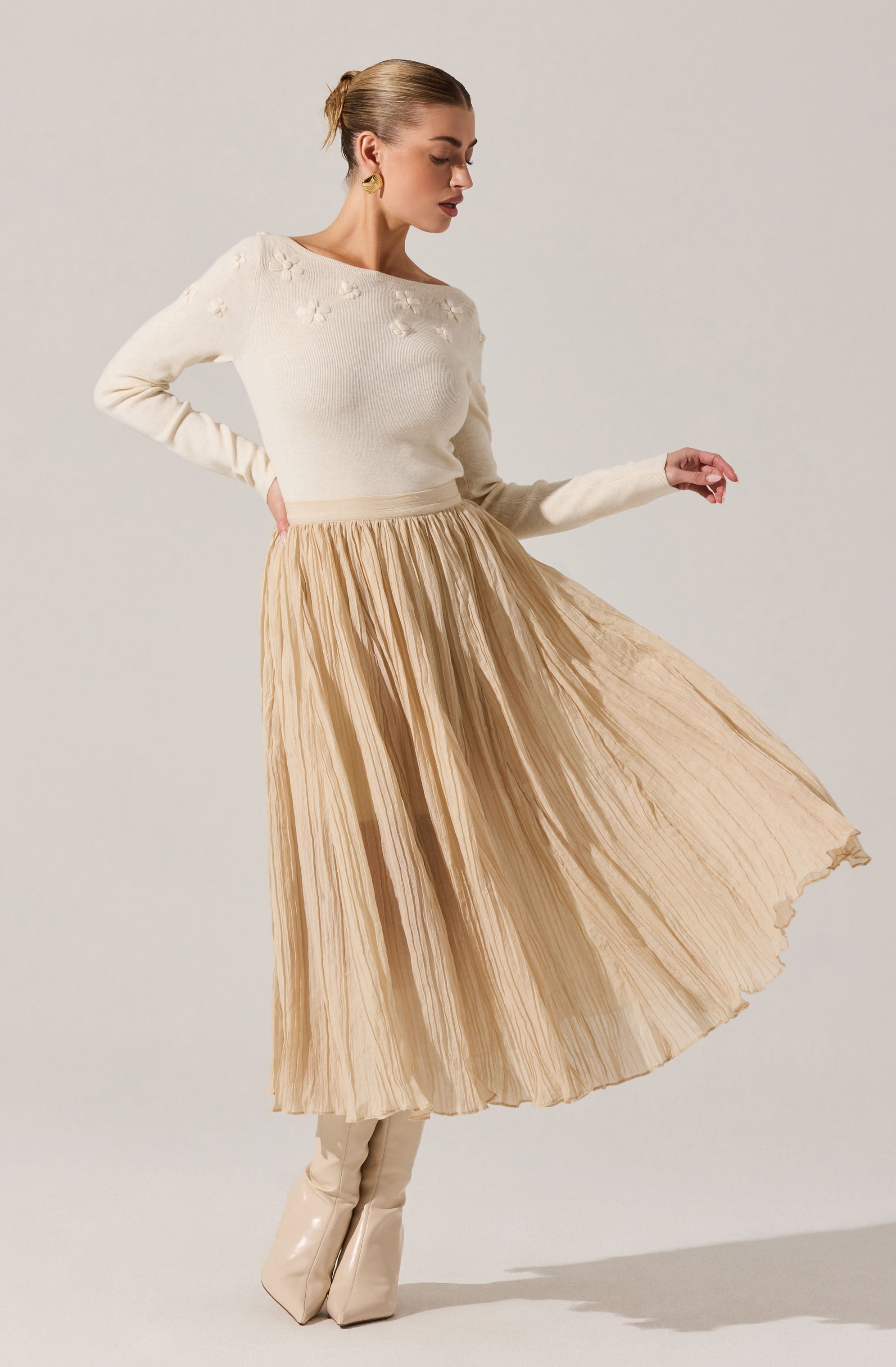 A woman is wearing a cream-colored sweater with floral embroidery on the chest and shoulders, paired with a high-waisted, pleated, beige midi skirt that flows as she moves. She completes the outfit with matching beige shiny boots and gold earrings.