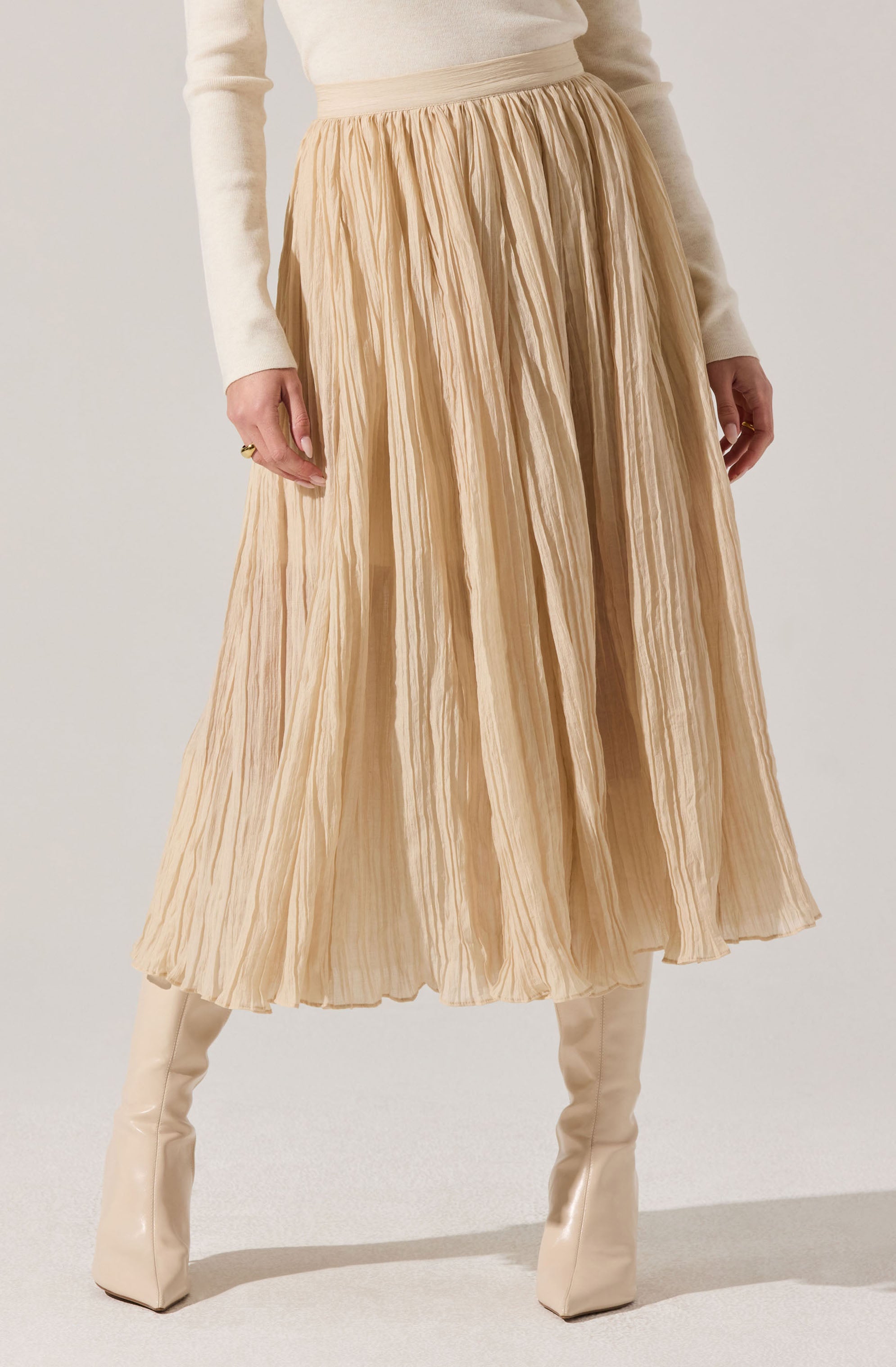 A person is wearing a light cream-colored long-sleeved top tucked into a high-waisted, pleated midi skirt made of a sheer, crinkled fabric in a soft beige tone. They are also wearing knee-high, shiny cream-colored boots.