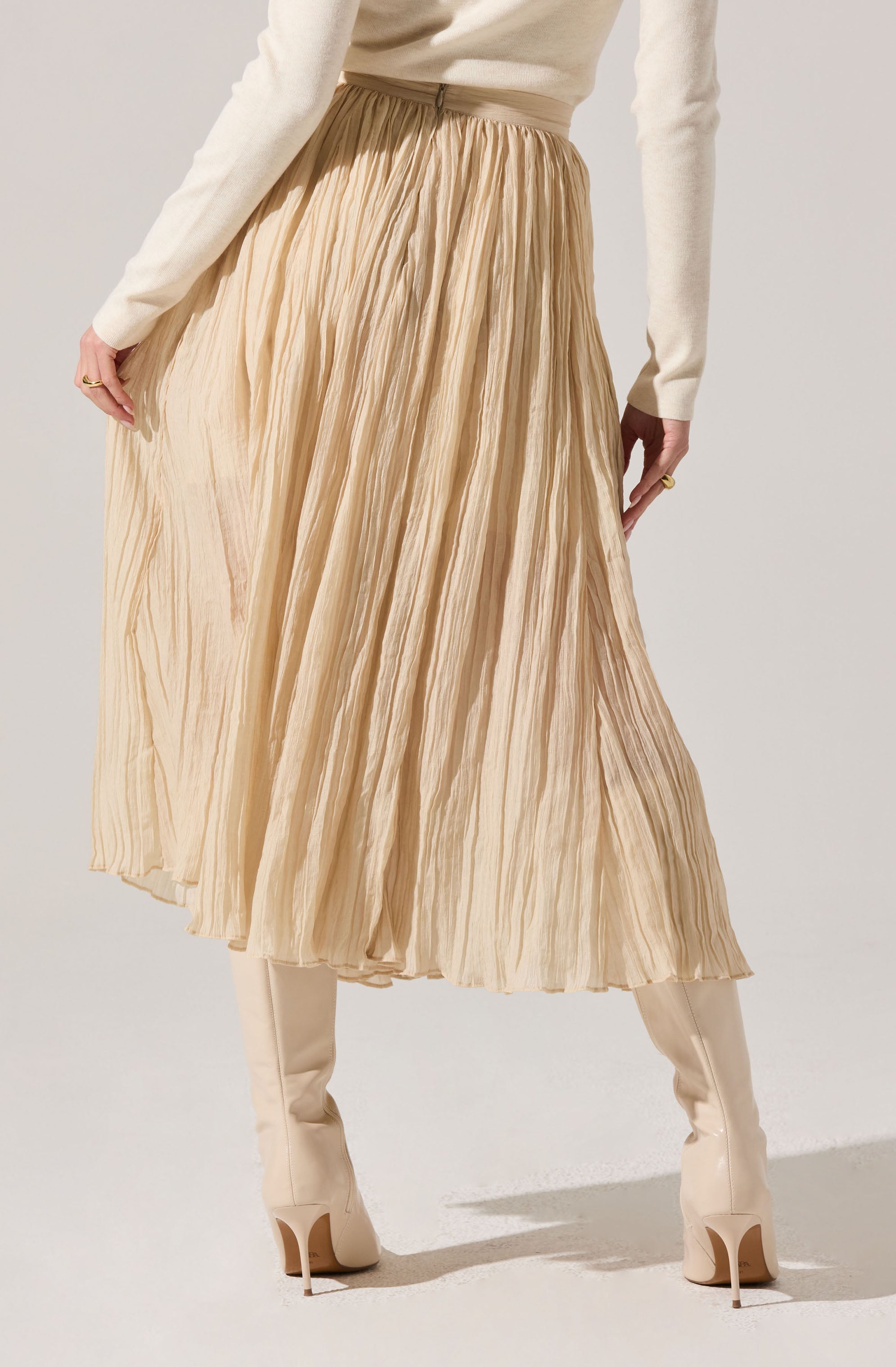 A woman is wearing a long, pleated, beige skirt with a high waist. She has paired it with knee-high, off-white boots with high heels. She is also wearing a long-sleeved, cream-colored top.