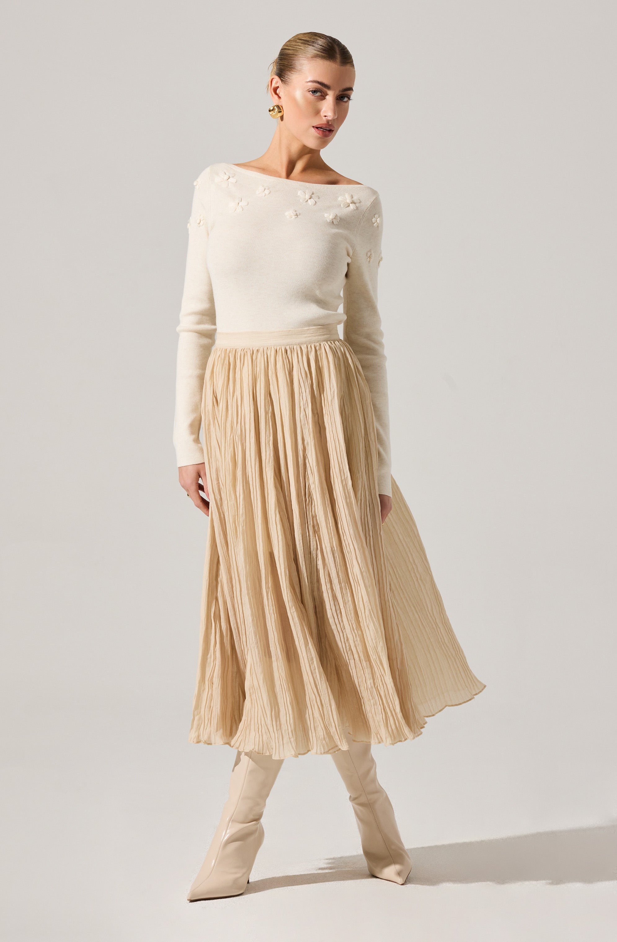 A woman is wearing a white long-sleeve sweater with embroidered floral details on the shoulders and upper chest. She pairs it with a beige, pleated, midi-length skirt that has a flowing, lightweight appearance. Her outfit is completed with knee-high beige boots, and she has her hair slicked back with large gold hoop earrings.
