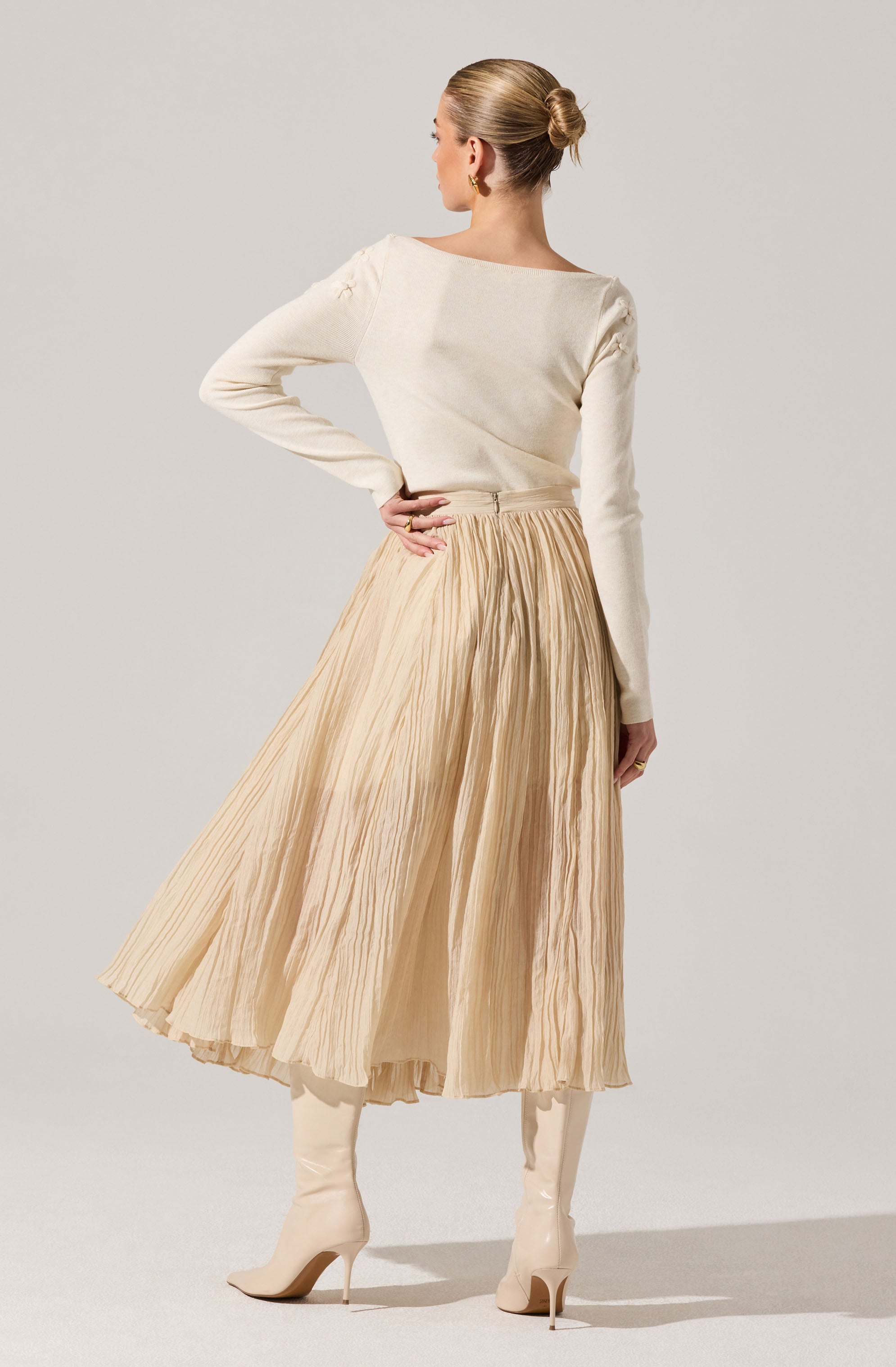 A woman is wearing a long-sleeved, cream-colored top and a high-waisted, beige pleated midi skirt. She has on knee-high, cream-colored heeled boots. Her hair is styled in a neat bun, and she is accessorized with gold earrings.