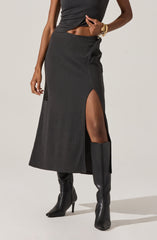 The outfit includes a black sleeveless crop top made of a textured fabric, paired with a matching high-waisted black midi skirt that features a thigh-high slit on the left side. The model is wearing tall black leather boots with high heels that extend above the knee. She is accessorized with gold jewelry, including bracelets on her left wrist.