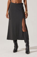 A woman is wearing a long, dark-colored wrap skirt with a high slit on one side, revealing her thigh and knee-high black leather boots. The skirt has a textured fabric and a knot detail at the waist. She is accessorized with gold bracelets and rings on her left arm and hand.