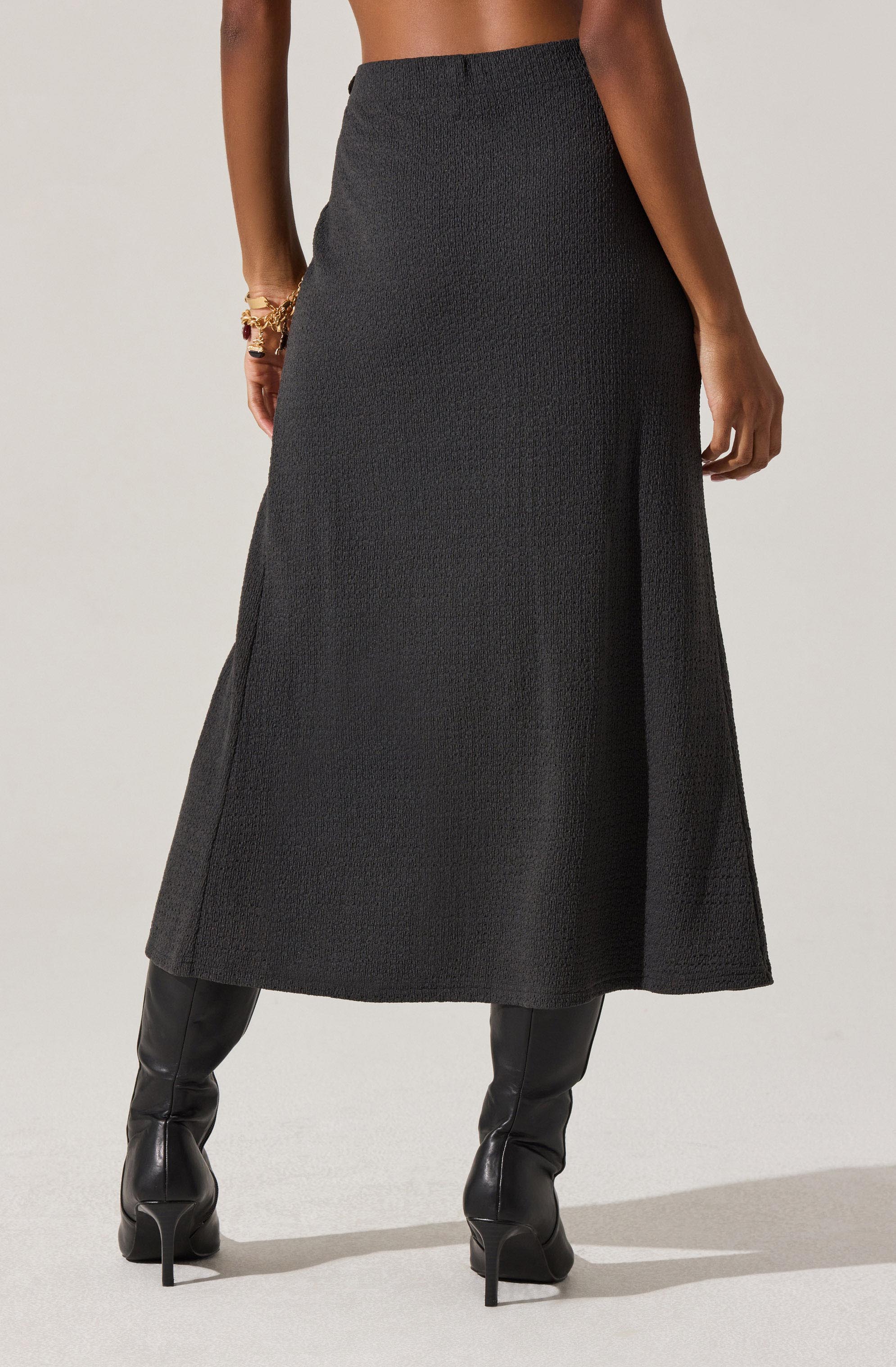 A woman is wearing a long, high-waisted, textured black skirt that reaches her mid-calf. She has on black, high-heeled leather boots. On her left wrist, she is wearing multiple gold bracelets.