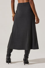 A woman is wearing a long, high-waisted, textured black skirt that reaches her mid-calf. She has on black, high-heeled leather boots. On her left wrist, she is wearing multiple gold bracelets.