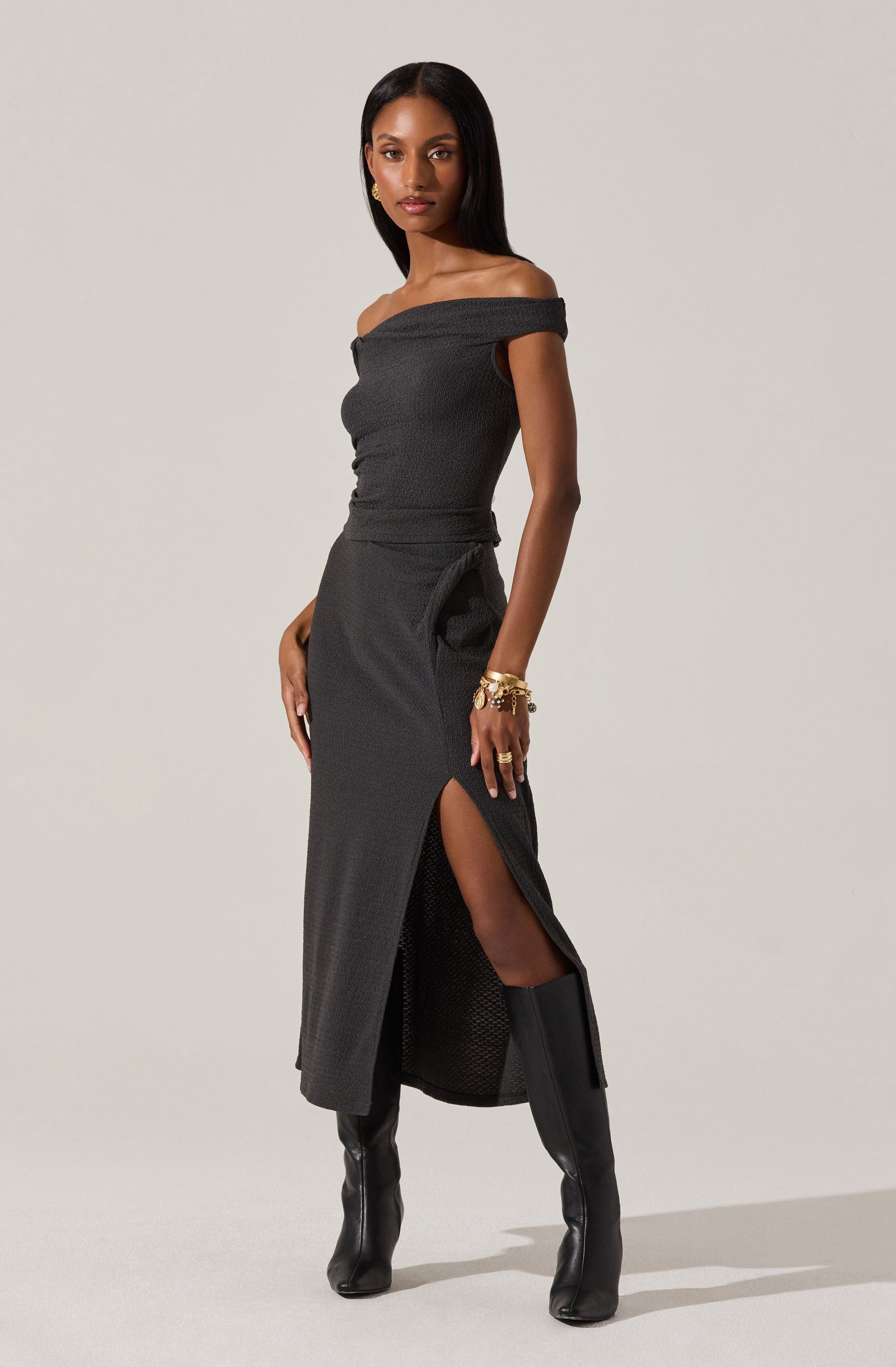 A woman is wearing a fitted, off-the-shoulder black dress with a textured fabric. The dress has a high slit on one side, revealing her leg and knee-high black leather boots. She accessorizes with multiple gold bracelets on her wrist.