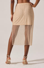 The image shows a woman wearing a beige skirt with a high waist and a fitted silhouette. The skirt has a dual-layer design: the top layer is solid and opaque, ending above the knee, while the bottom layer is made of sheer, see-through fabric extending to mid-calf length. The sheer portion has a dotted pattern, creating a semi-transparent effect. She is also wearing beige high-heeled sandals with ankle straps.
