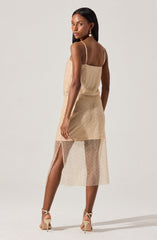 A woman is wearing a sleeveless, beige dress with thin spaghetti straps. The dress has a layered design, featuring a solid upper layer and a see-through, dotted lower layer that extends below the knee. There is a slit on the side of the lower layer, revealing her leg. She is also wearing high-heeled shoes and statement earrings.