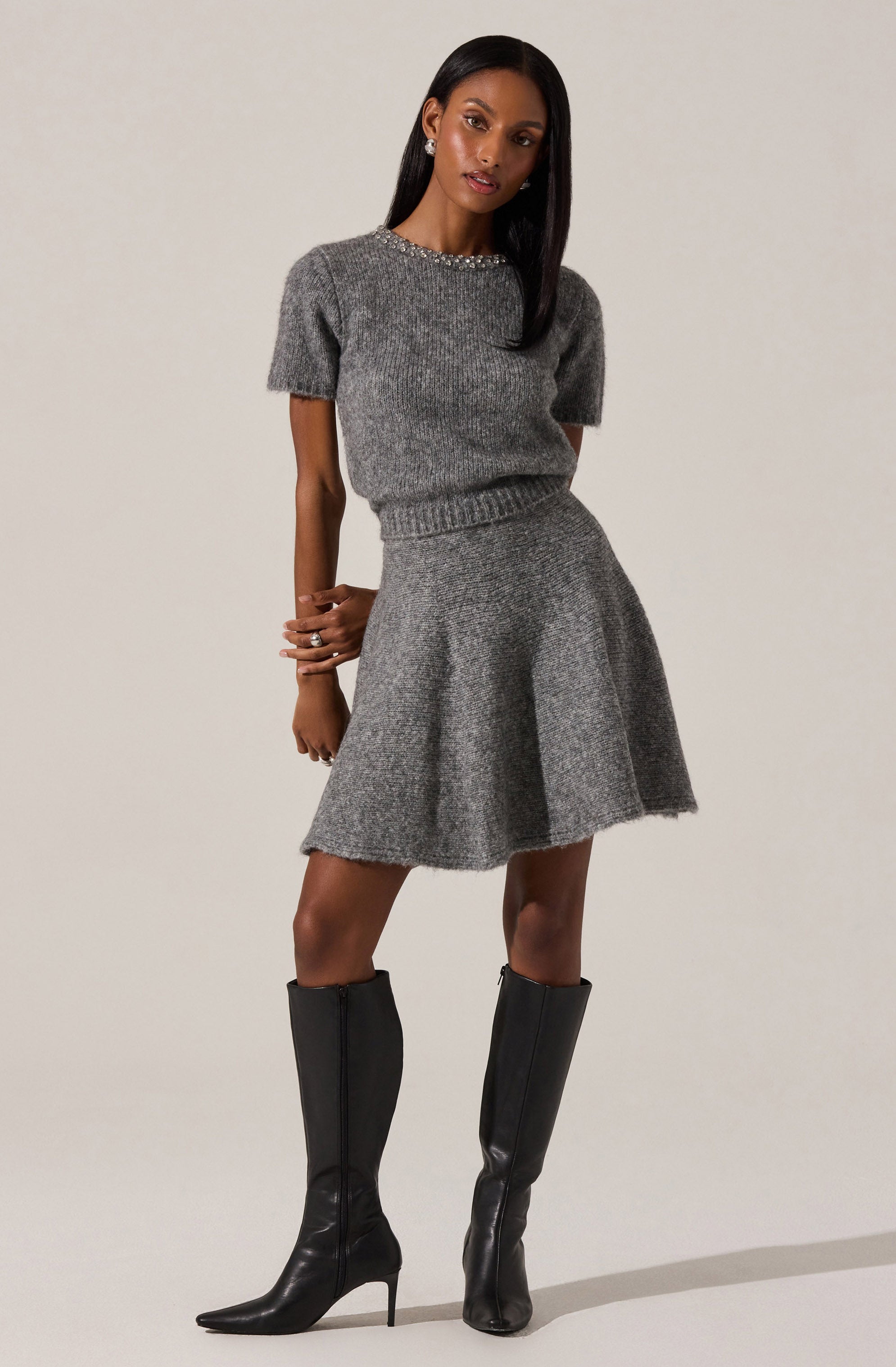 The woman is wearing a matching gray knit sweater and skirt set. The sweater has short sleeves and a round neckline decorated with a row of embellishments. The skirt is flared and falls above the knee. She is also wearing tall black leather boots with high heels.