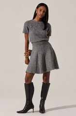 The woman is wearing a matching gray knit sweater and skirt set. The sweater has short sleeves and a round neckline decorated with a row of embellishments. The skirt is flared and falls above the knee. She is also wearing tall black leather boots with high heels.