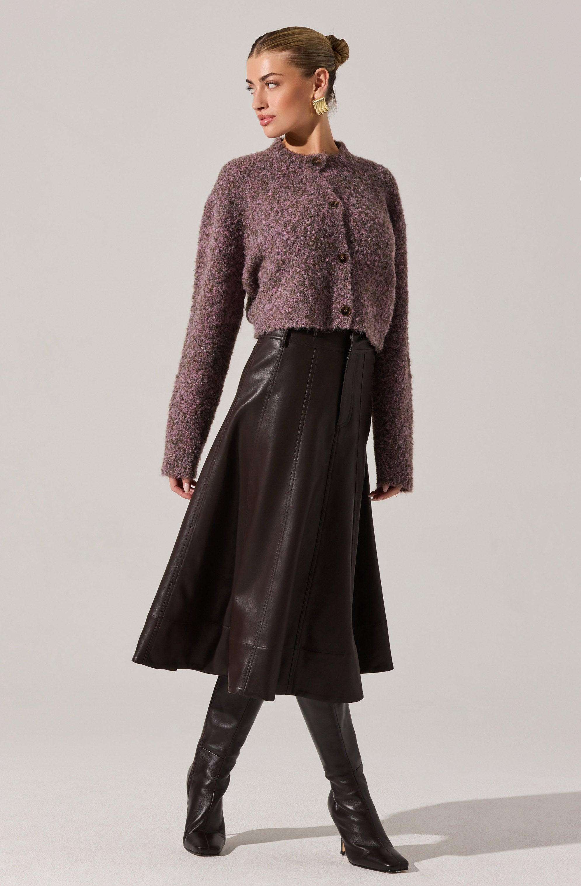 A woman is wearing a textured, cropped, long-sleeve cardigan in a purple and gray mix, with a rounded neckline and buttons down the front. She pairs it with a high-waisted, dark brown, leather midi skirt that flares out slightly, and matching thigh-high leather boots. She is accessorized with large, gold hoop earrings and has her hair styled in a sleek, low bun.