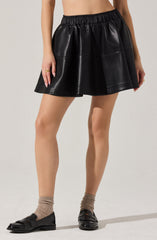A person is wearing a black faux leather skirt with an elastic waistband, featuring a slight flare and shiny finish. They are also wearing taupe socks and black shiny loafers with fringe details. The person's upper body is not visible, and there are no additional accessories or clothing items shown.