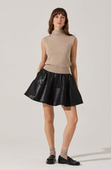 A woman is wearing a sleeveless beige turtleneck sweater and a black faux leather flared skirt. She has on beige socks and black loafers.