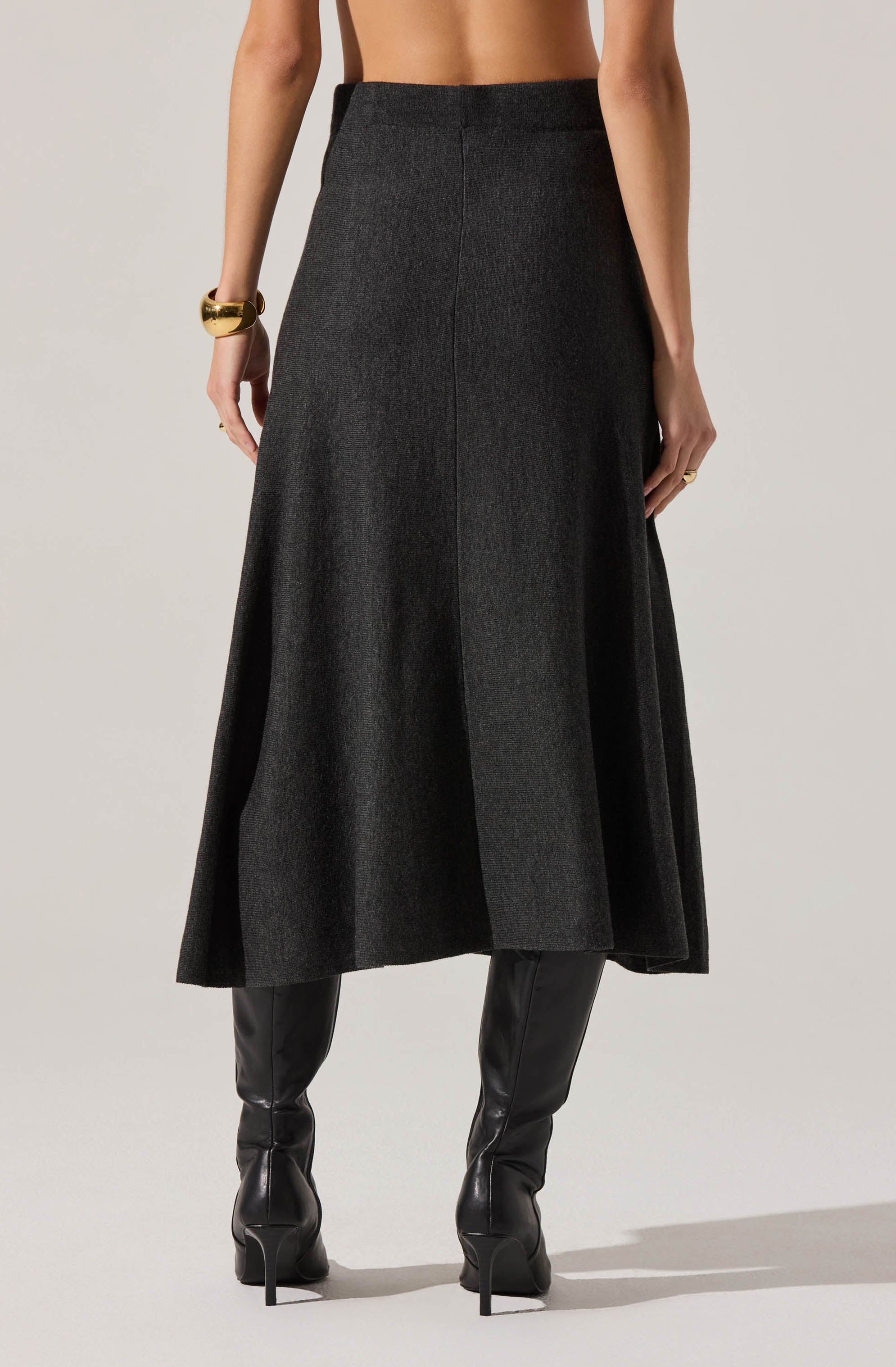 A person is wearing a high-waisted, long, dark gray skirt with a subtle texture. The skirt has a simple, clean design with a front seam running vertically down the center. They are also wearing black, knee-high leather boots with pointed toes and high heels. The person has accessorized with a chunky gold bracelet on their left wrist and a gold ring on their right hand. The background is plain and light-colored.