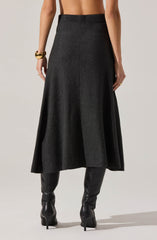 A person is wearing a high-waisted, long, dark gray skirt with a subtle texture. The skirt has a simple, clean design with a front seam running vertically down the center. They are also wearing black, knee-high leather boots with pointed toes and high heels. The person has accessorized with a chunky gold bracelet on their left wrist and a gold ring on their right hand. The background is plain and light-colored.