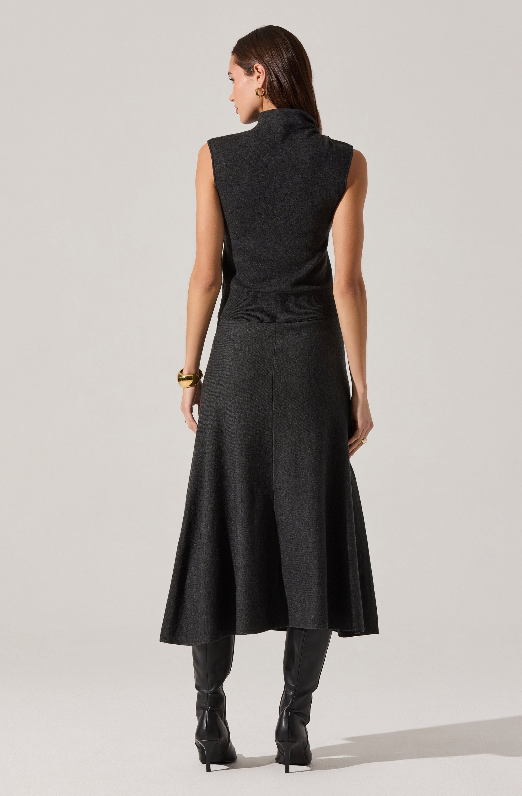 A woman is wearing a sleeveless, high-neck, dark gray knit top paired with a long, flowy, dark gray skirt. She has gold earrings, a gold bracelet on her right wrist, and black high-heeled boots.
