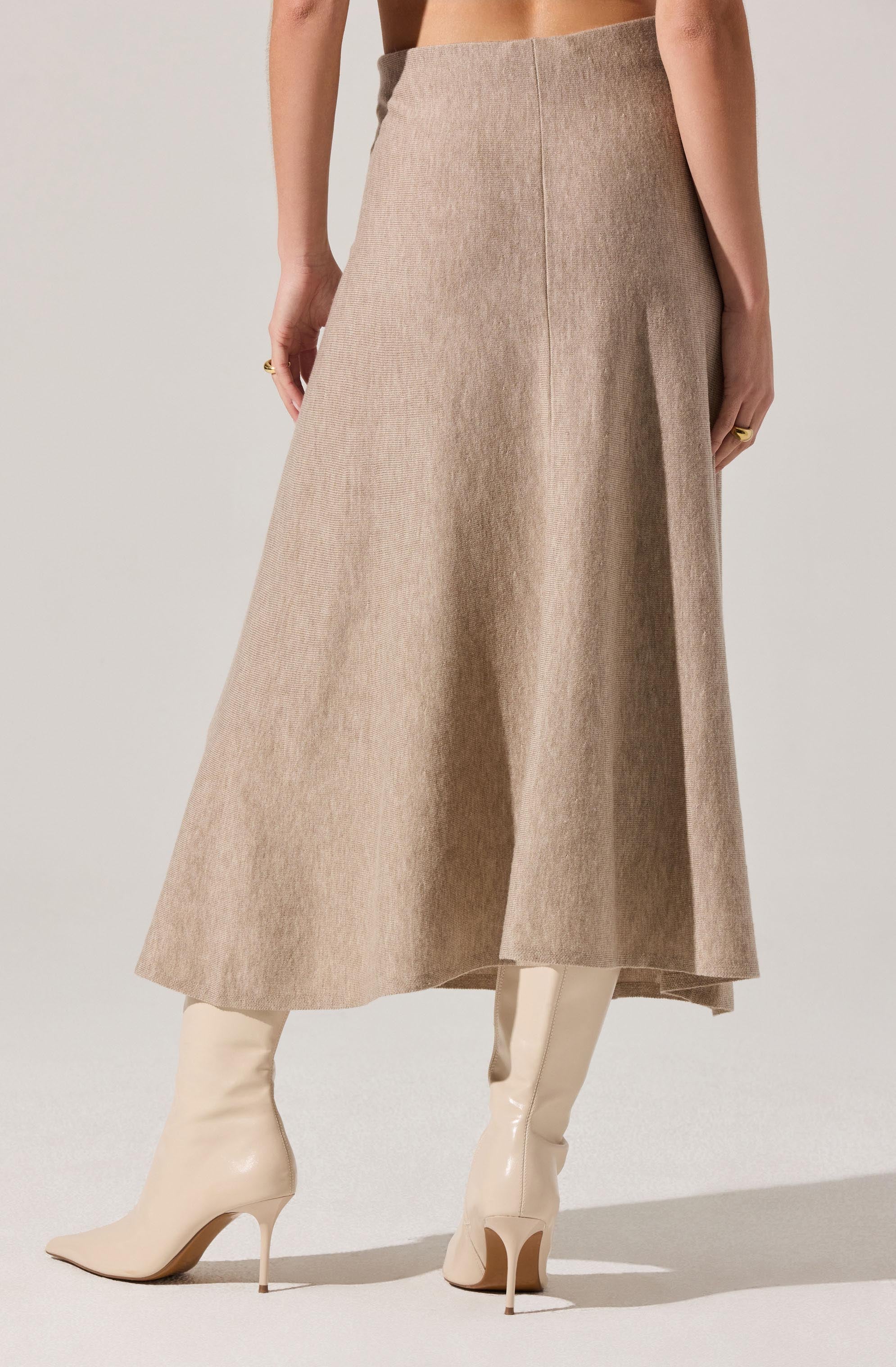 A woman is wearing a high-waisted, ankle-length beige skirt with a subtle texture and a front center seam. She pairs it with white, knee-high, glossy stiletto boots. The image captures her from the waist down, with no top or additional accessories visible.
