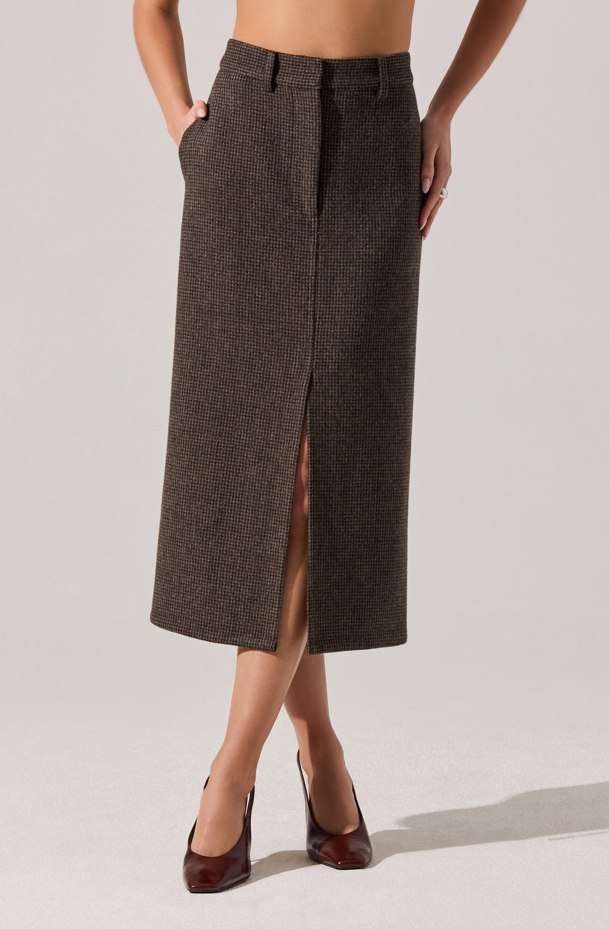 A woman is wearing a high-waisted, ankle-length skirt with a small checkered pattern in brown tones. The skirt features a front slit that extends from the hem to just above the knee. She has her hands in her pockets and is wearing dark burgundy high heels.