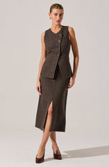 A woman is wearing a sleeveless, fitted brown vest with a subtle checkered pattern, featuring a small collar and three buttons on the front. She pairs it with a matching brown checkered skirt that reaches mid-calf and has a front slit. She completes the outfit with dark brown high-heeled shoes.