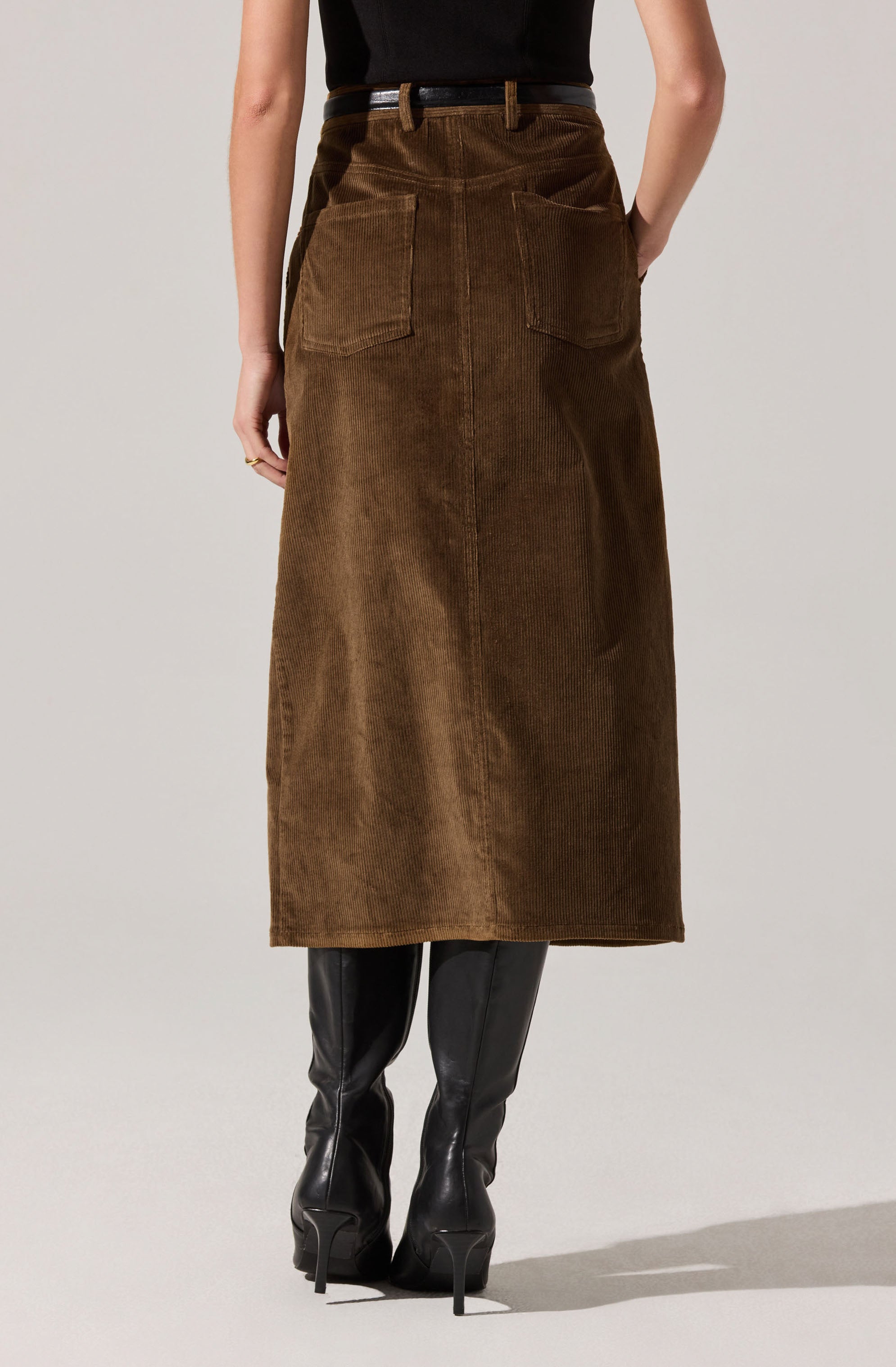 A person is wearing a high-waisted, long brown corduroy skirt with large back patch pockets. They have paired it with knee-high black leather boots with pointed toes and stiletto heels, and a black top tucked into the skirt.